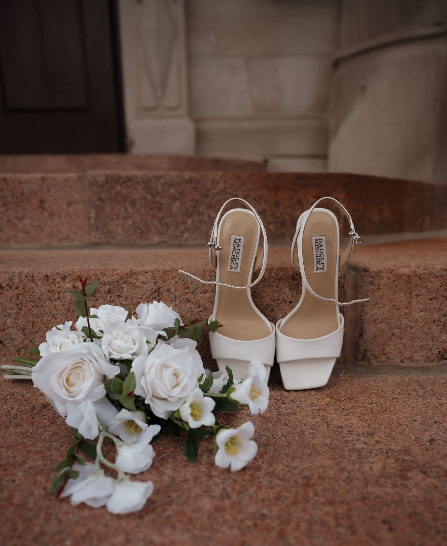 White bridal heels and a bouquet of white roses on stone steps outside a church.