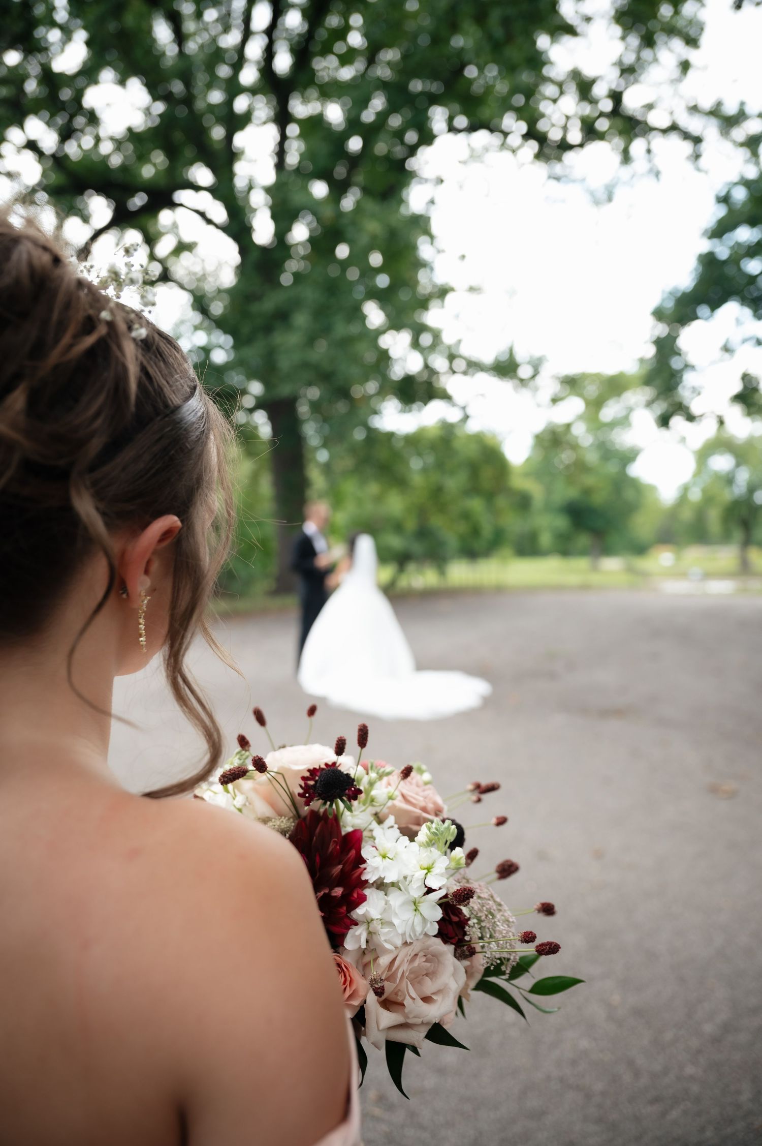 Bridesmaid holding a burgundy and blush floral bouquet while a bride and groom stand on a tree-lined path in the background.