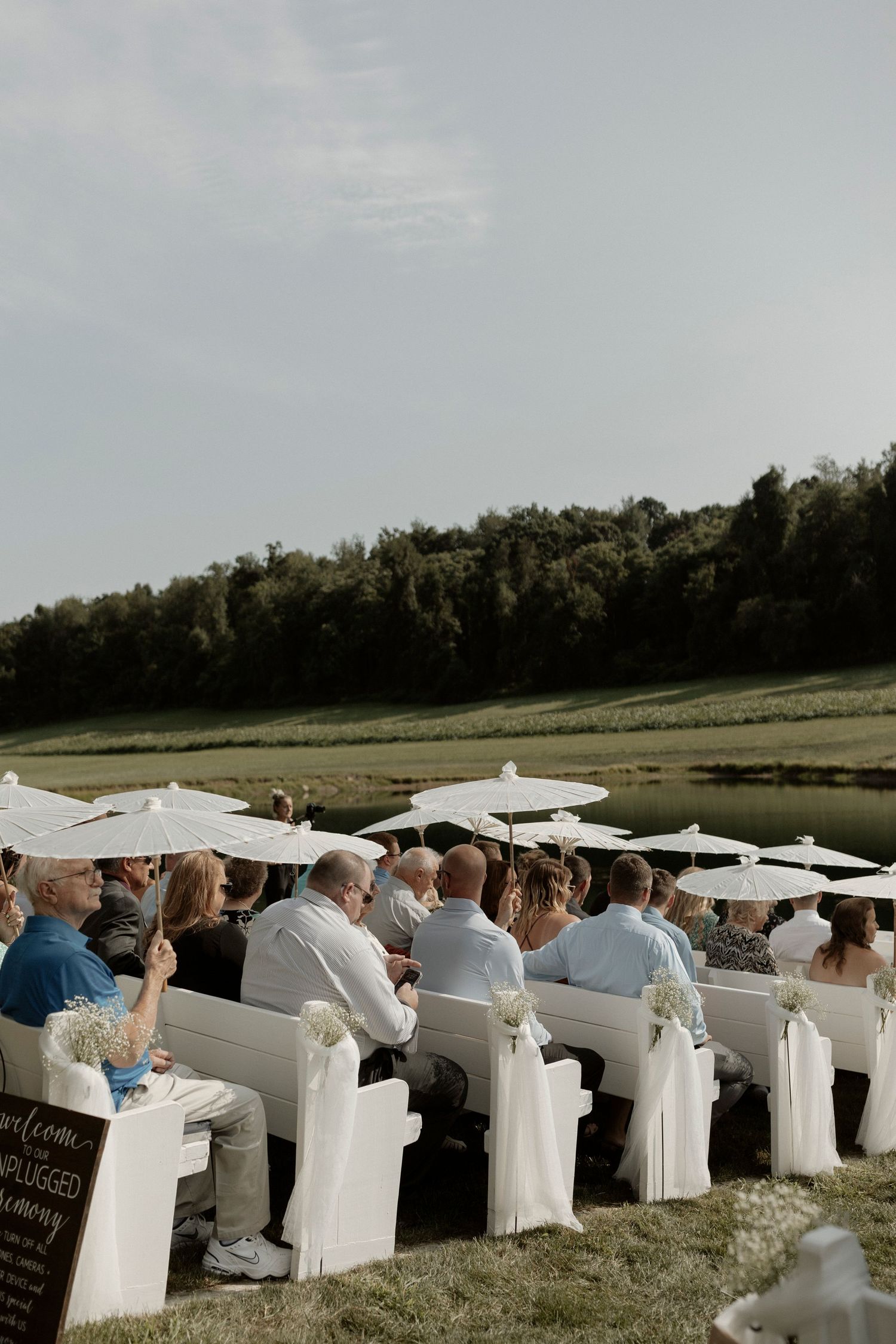 Wedding guests seated in white chairs with umbrellas at an outdoor lakeside ceremony on a grassy lawn.