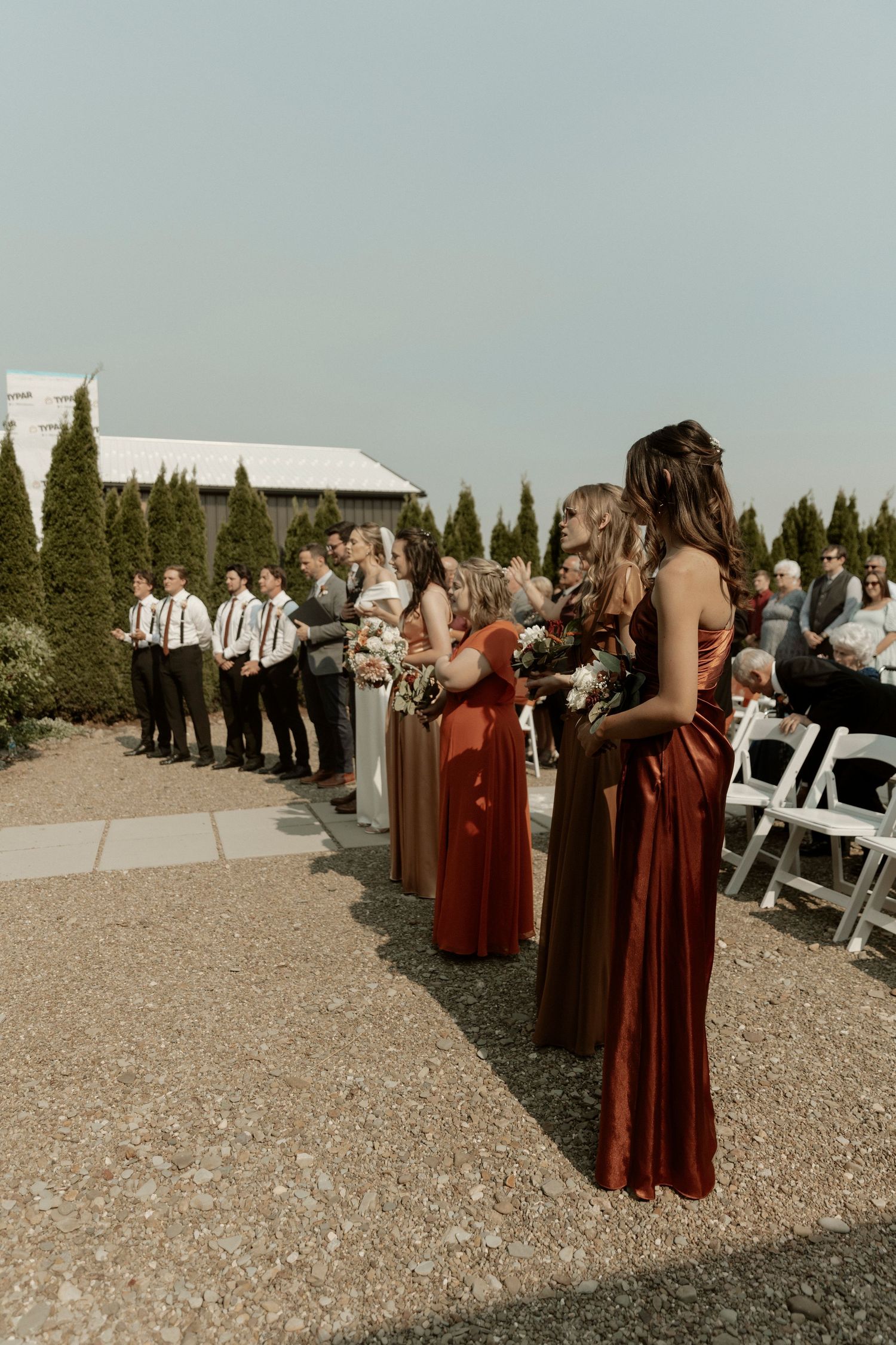 Wedding party members in burgundy dresses stand in line outdoors next to cypress trees during outdoor ceremony.