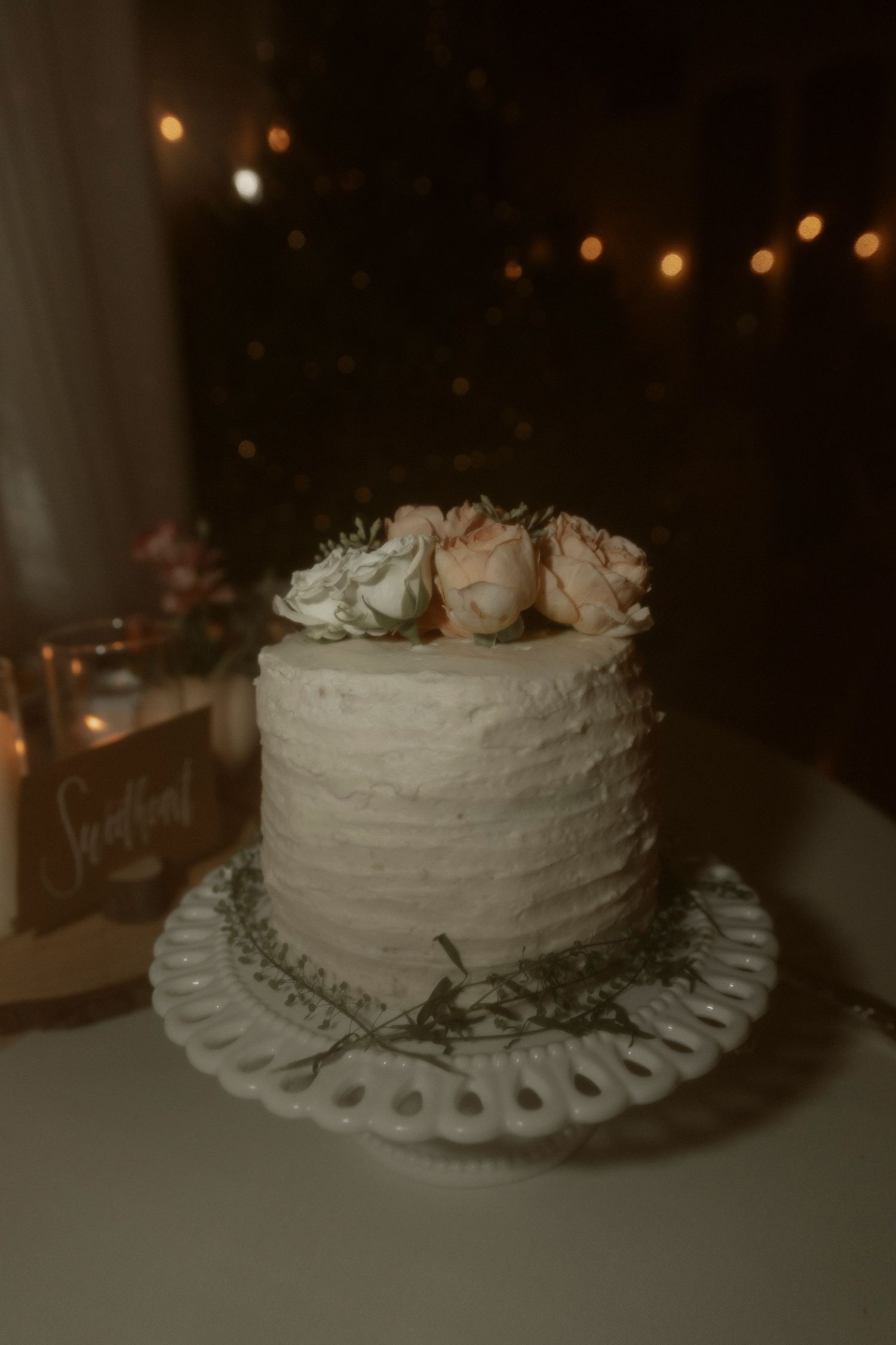 A white rustic wedding cake with roses on top sits on a decorative plate with blurred string lights in the background.