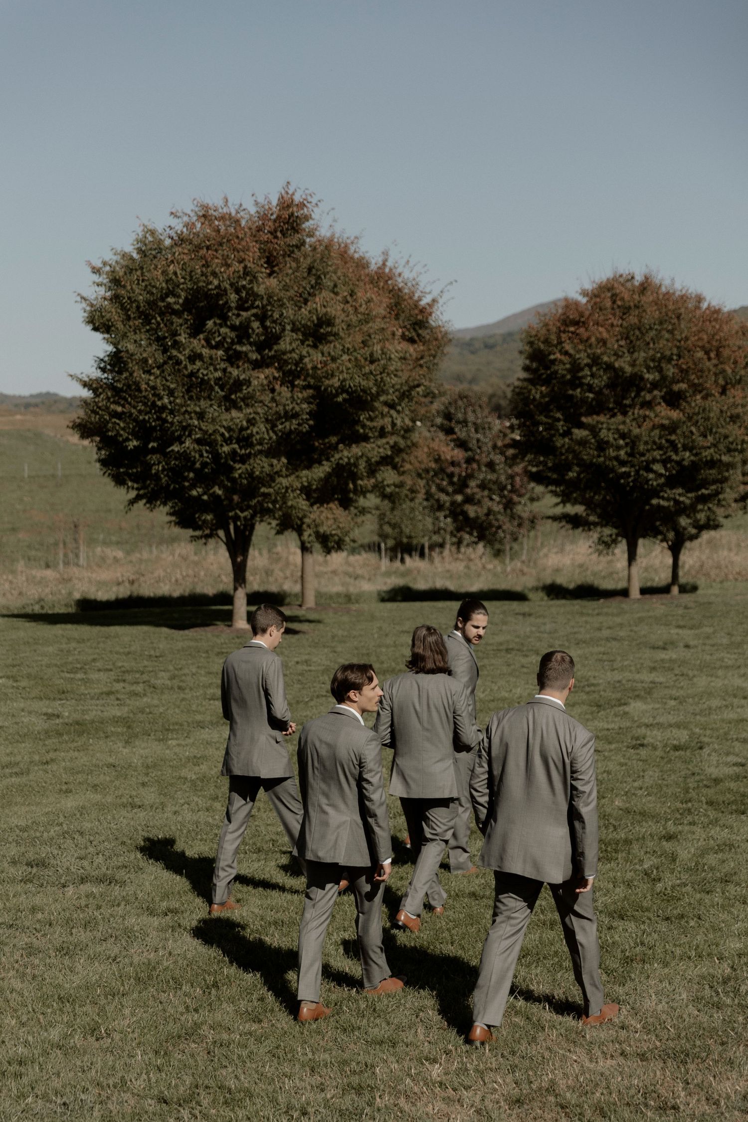Groomsmen in matching gray suits walking together in a grassy field with autumn trees in the background.