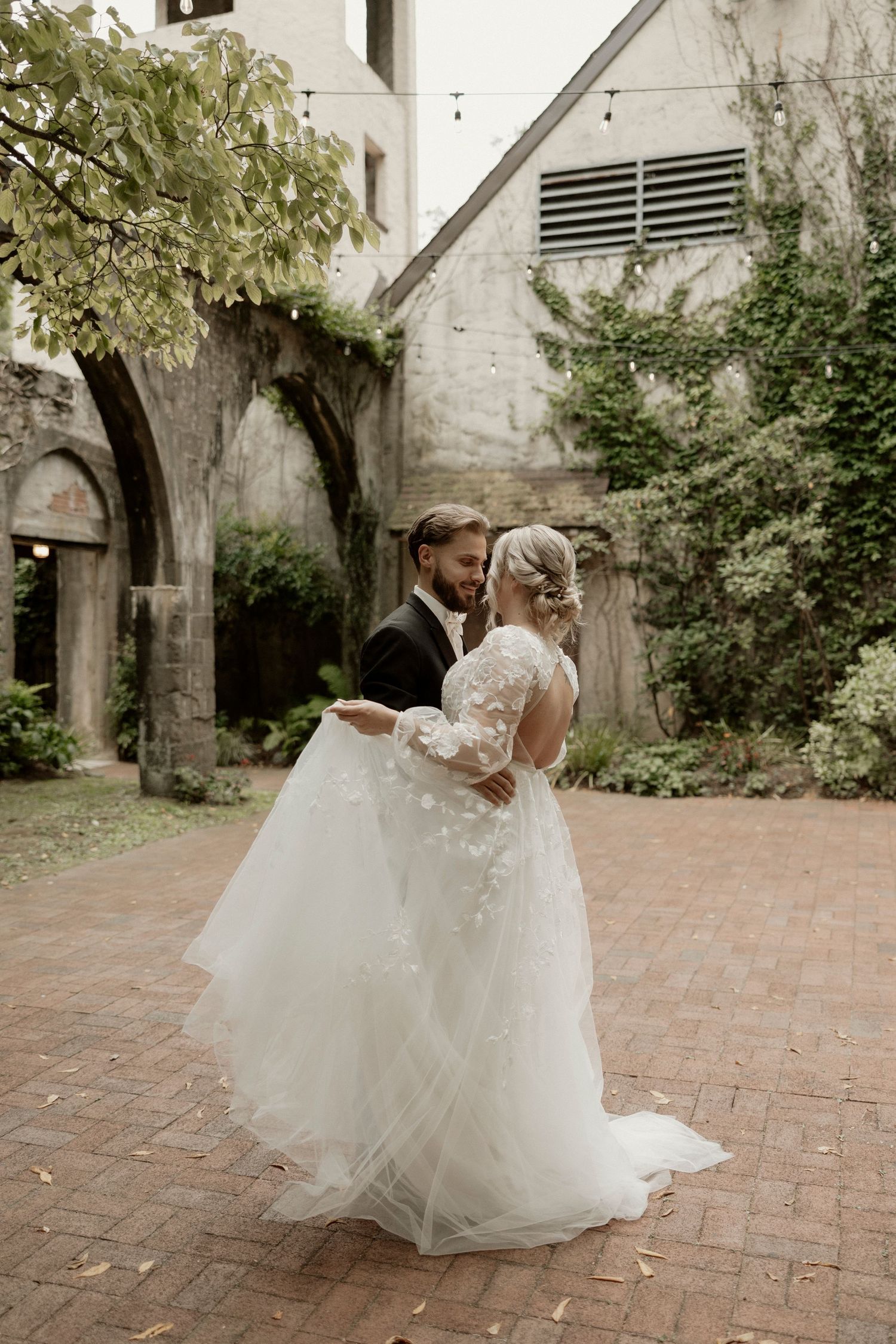 Newlywed couple dancing together in a courtyard with white wedding dress and lace details.