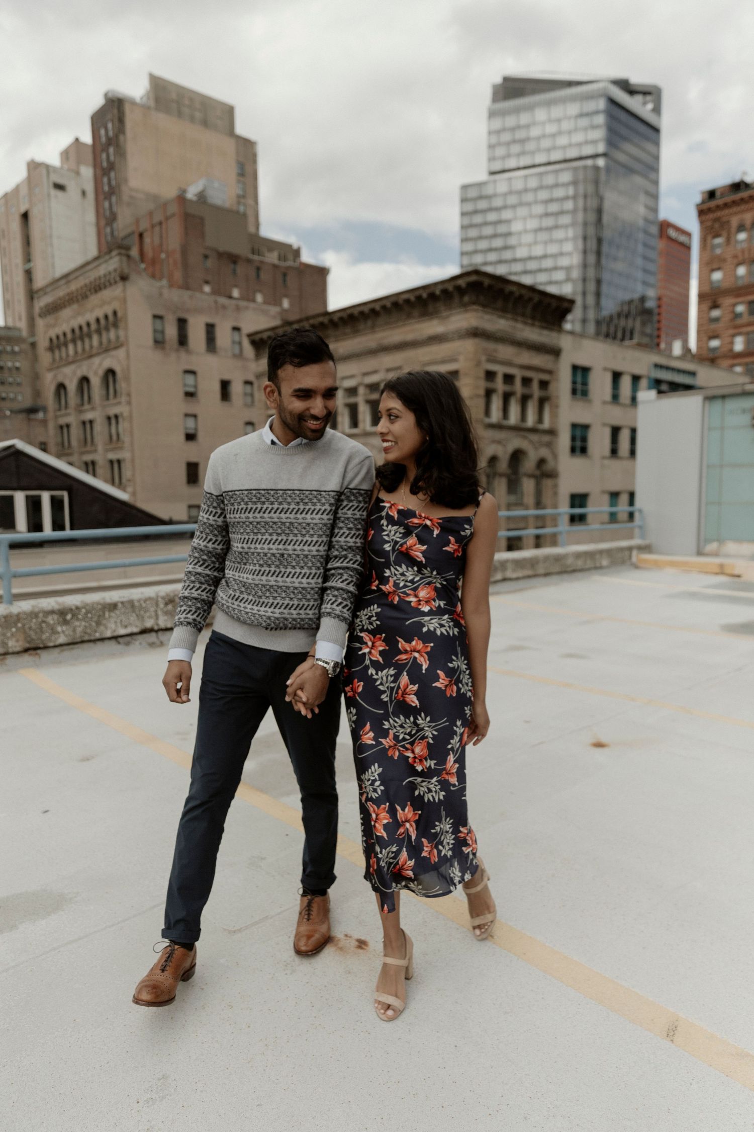 A couple holds hands on a rooftop in a city, wearing stylish outfits against an urban backdrop of tall buildings.