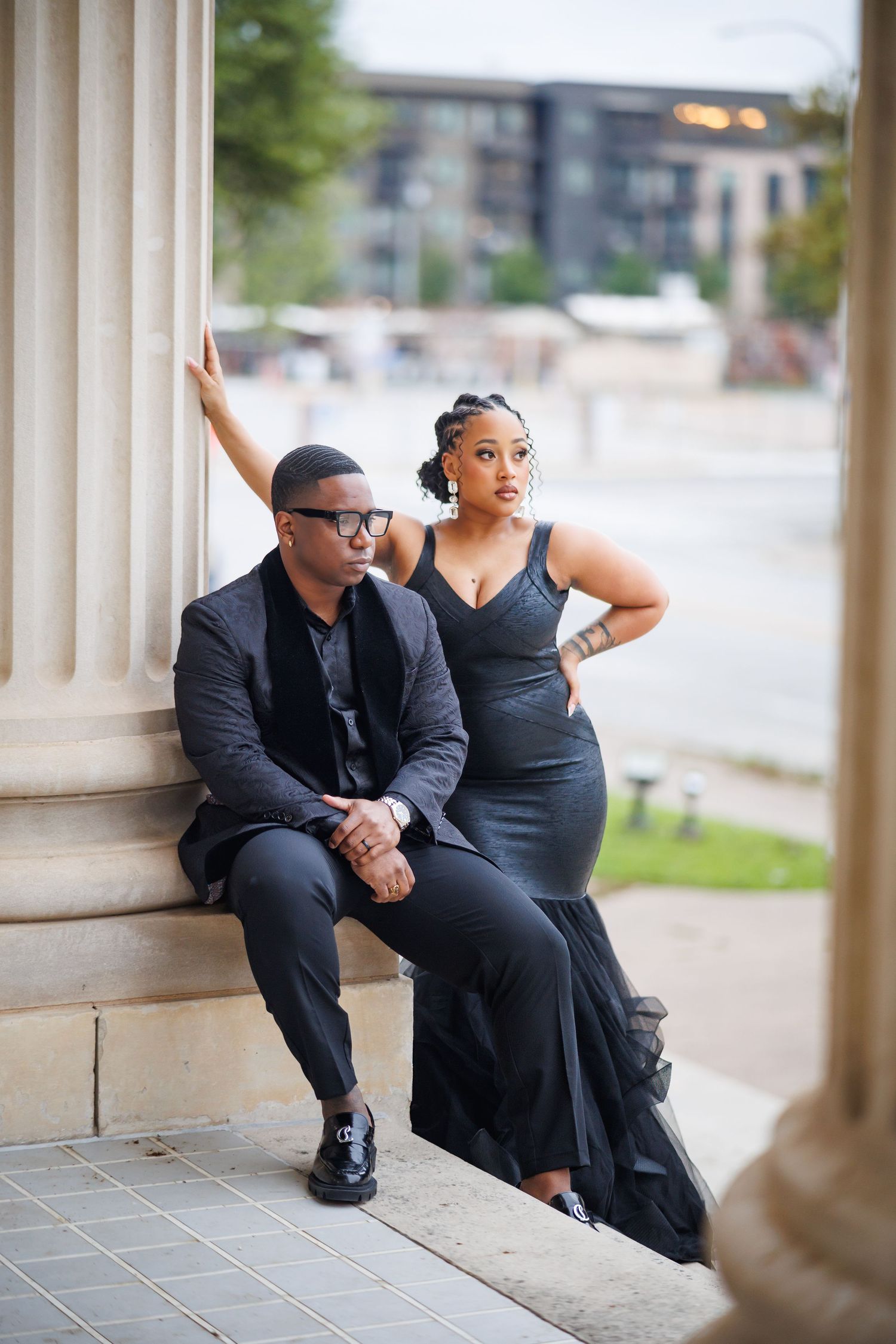A couple in formal black attire poses together on stone steps for an elegant photoshoot.