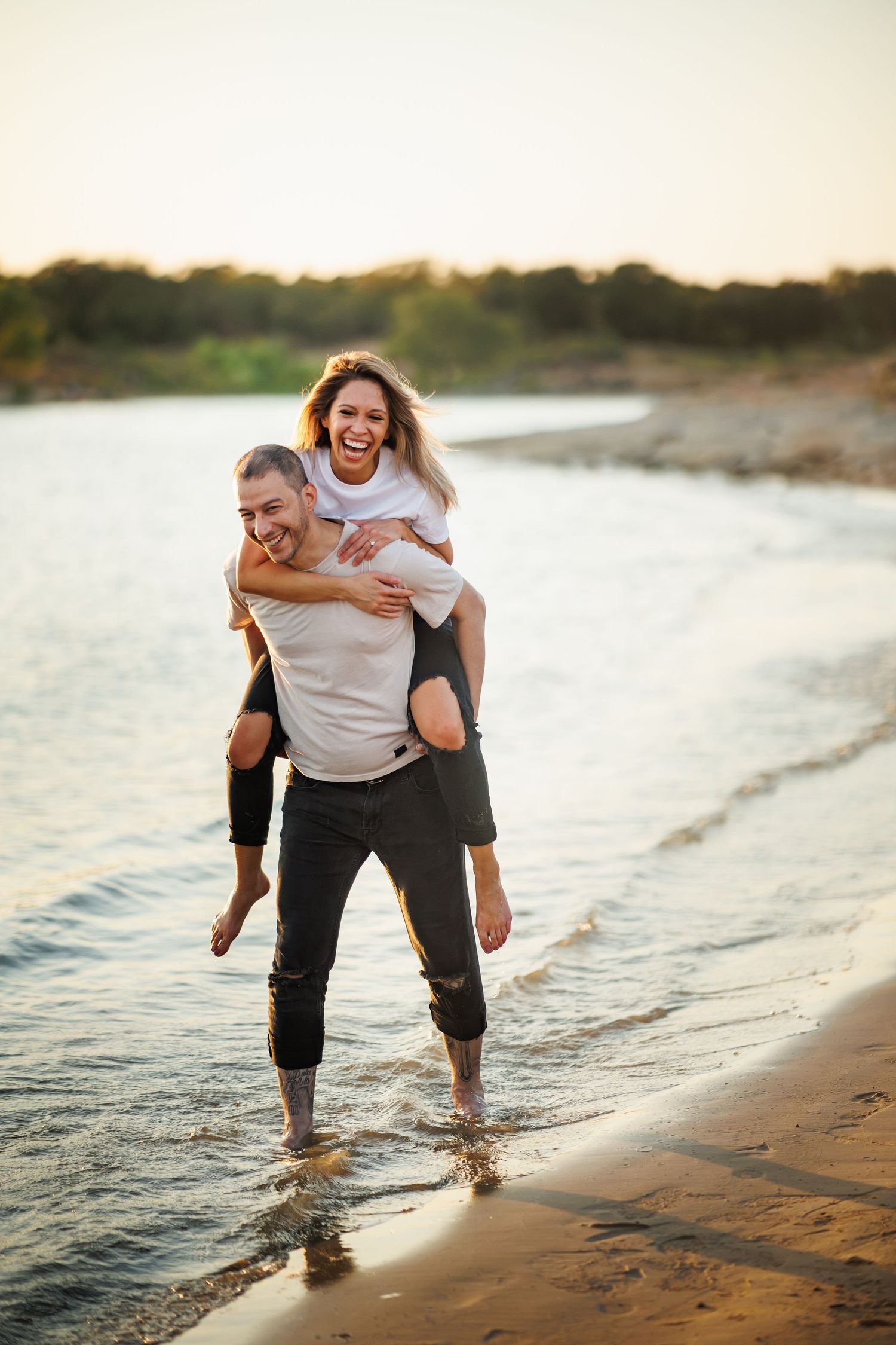 A couple enjoys a playful piggyback ride along a sandy beach at sunset with waves crashing nearby.