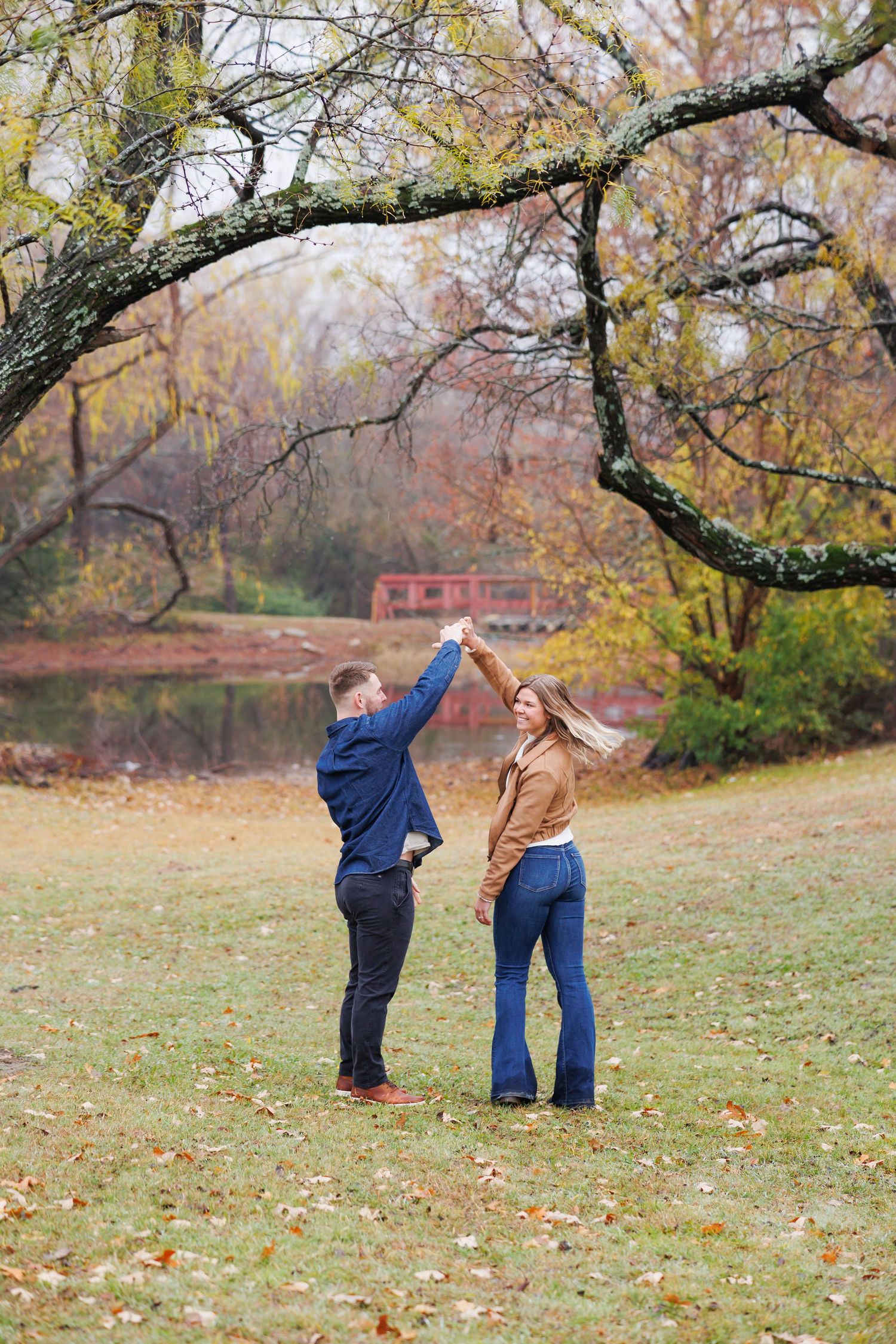 Couple dancing outdoors in autumn park near pond, woman in brown jacket and flare jeans, man in blue sweater.