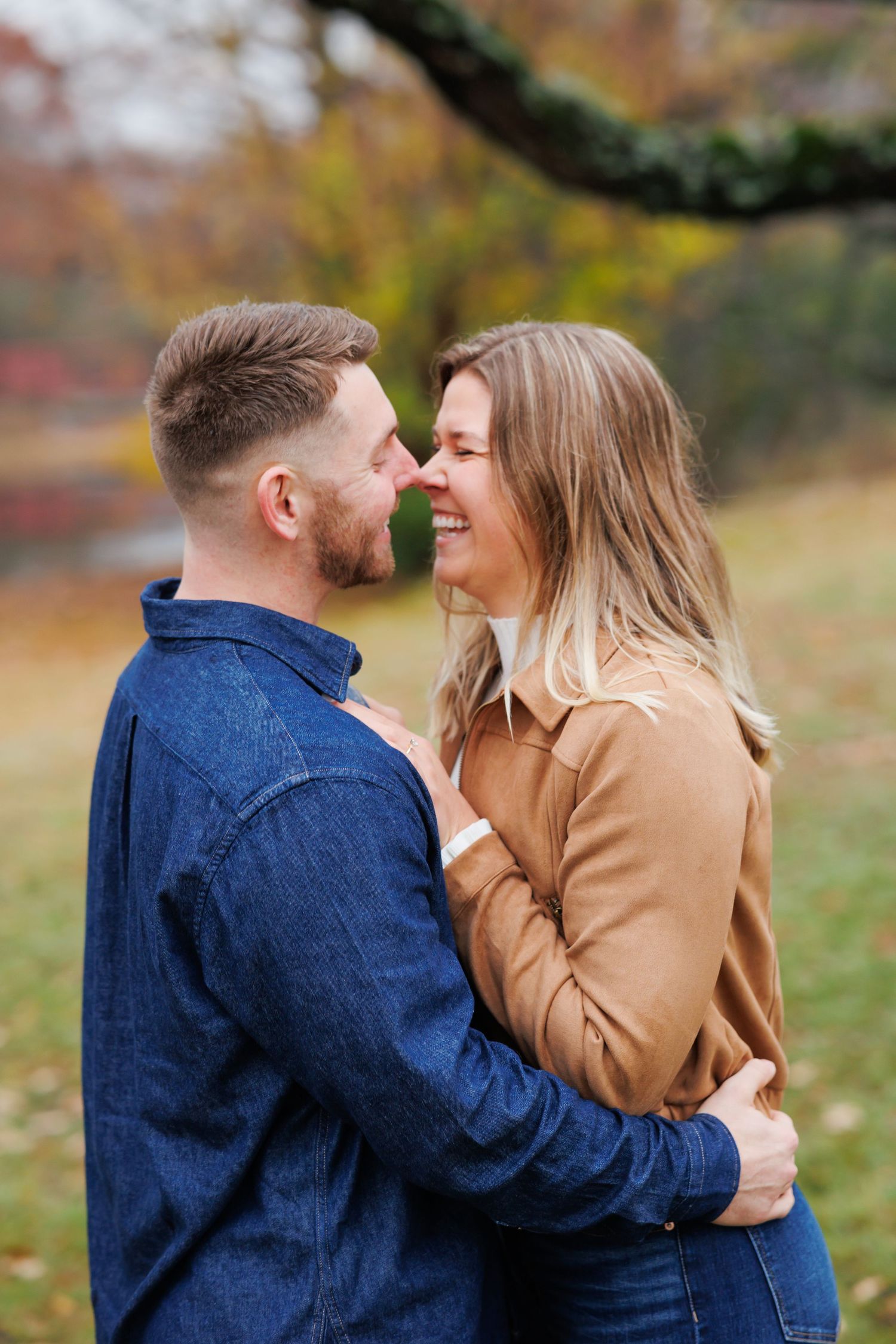 A smiling couple embraces outdoors in autumn, wearing denim and tan shirts, sharing a joyful moment among fall foliage.