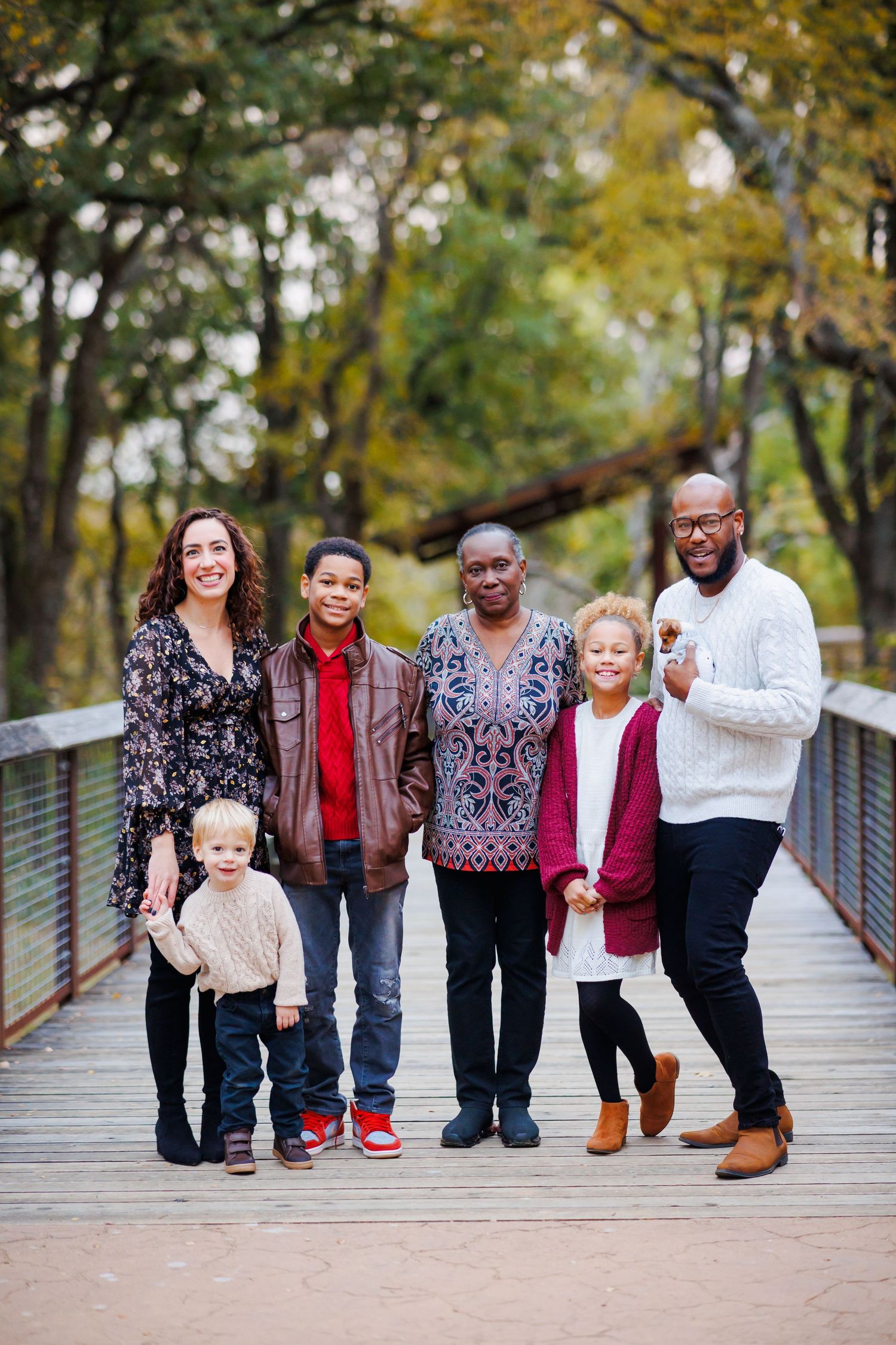 Multigenerational family of six posing together on a wooden bridge surrounded by autumn foliage in a park.