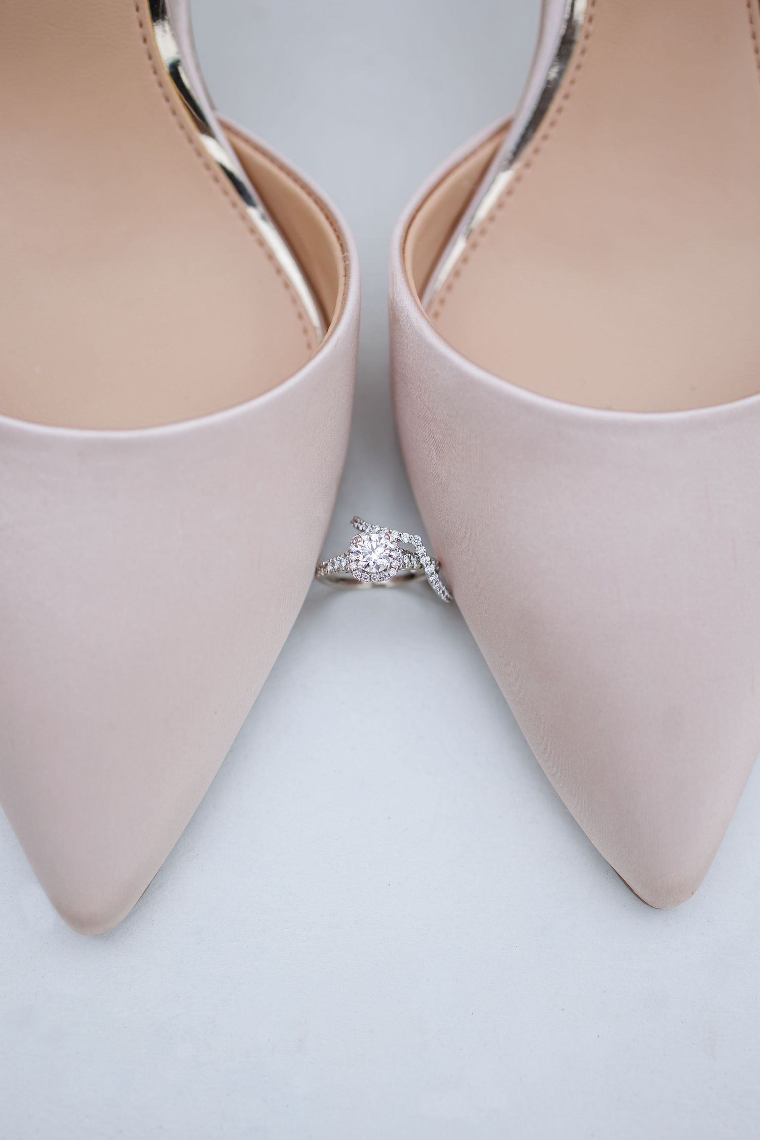 Diamond engagement ring positioned between blush pink bridal shoes on white surface.