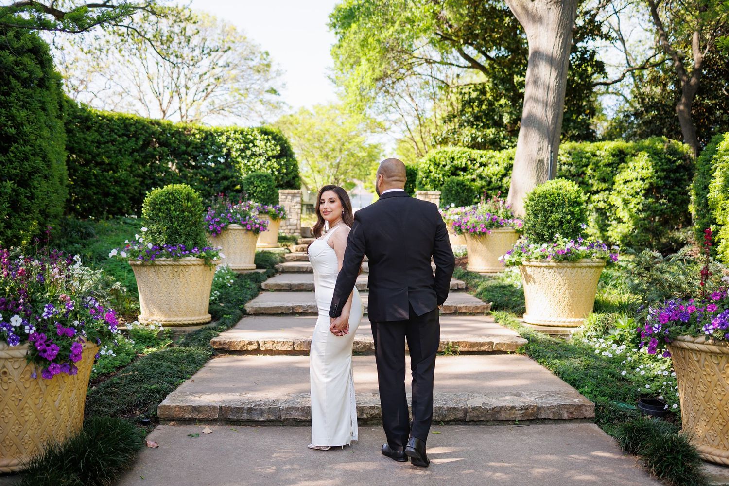 A couple walks up stone garden steps lined with ornate planters and purple flowers in a lush outdoor setting.