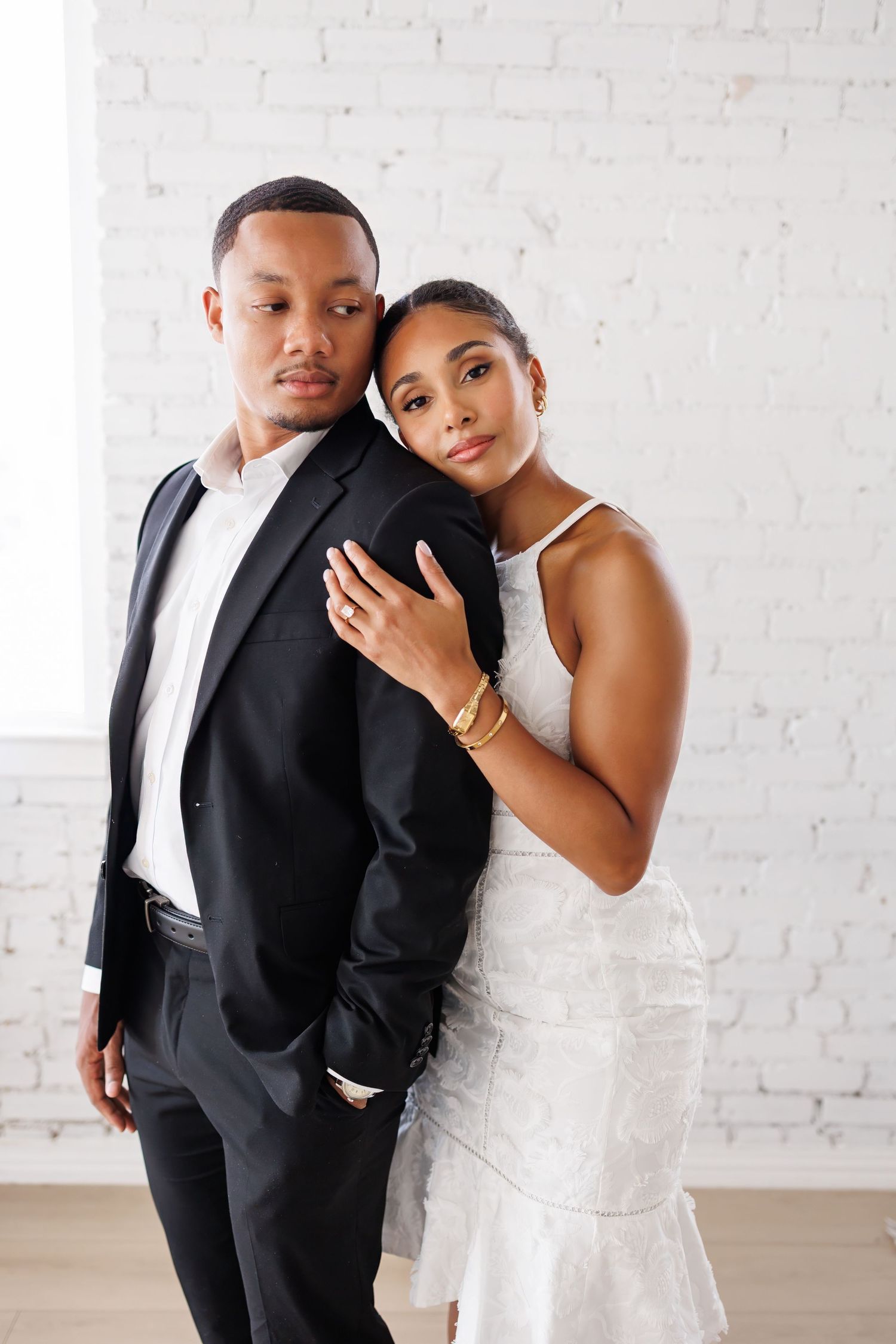 Elegant couple in formal wedding attire pose together against white brick wall in romantic studio portraits.