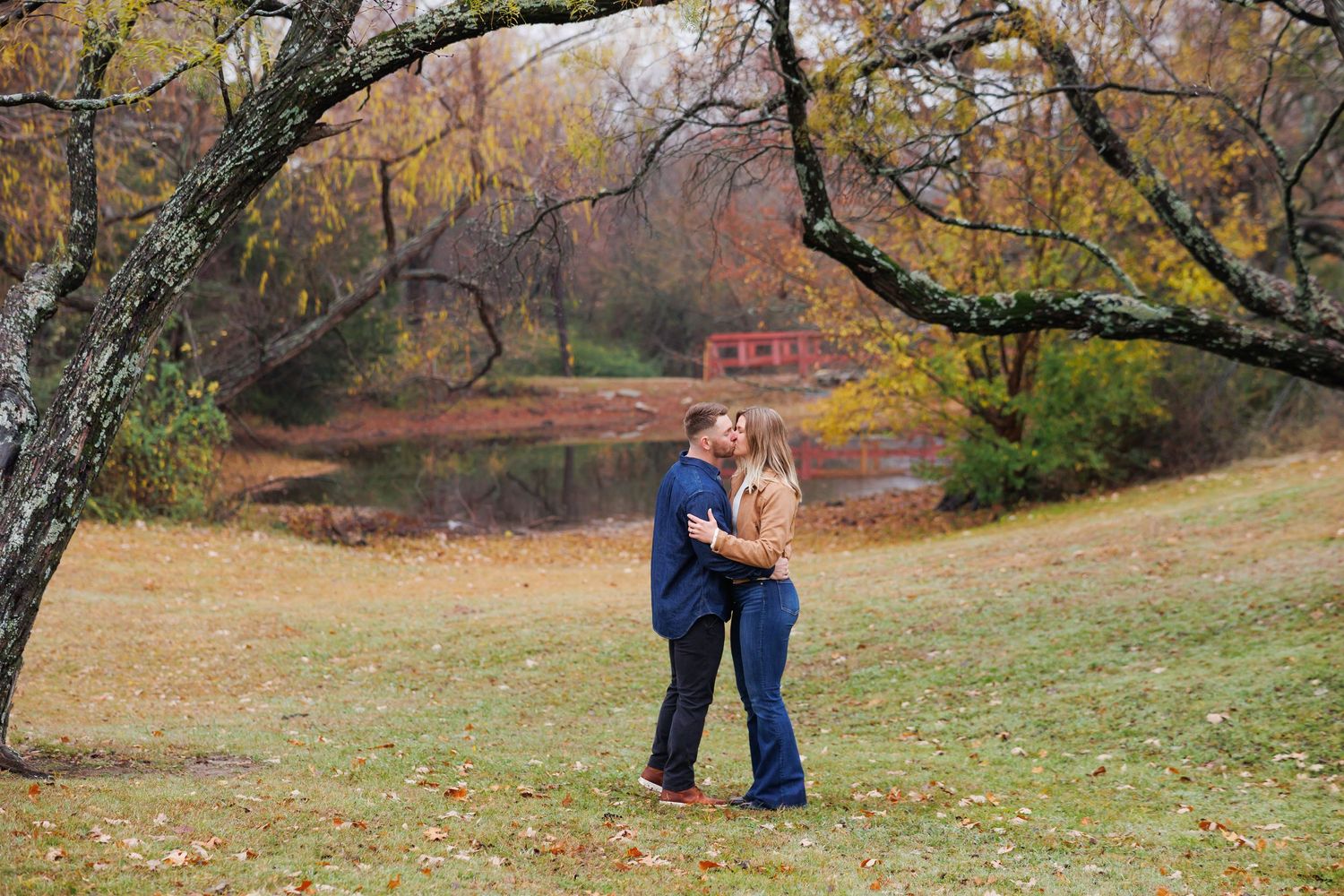 A couple shares a romantic moment in a park surrounded by autumn trees with golden leaves falling in the background.