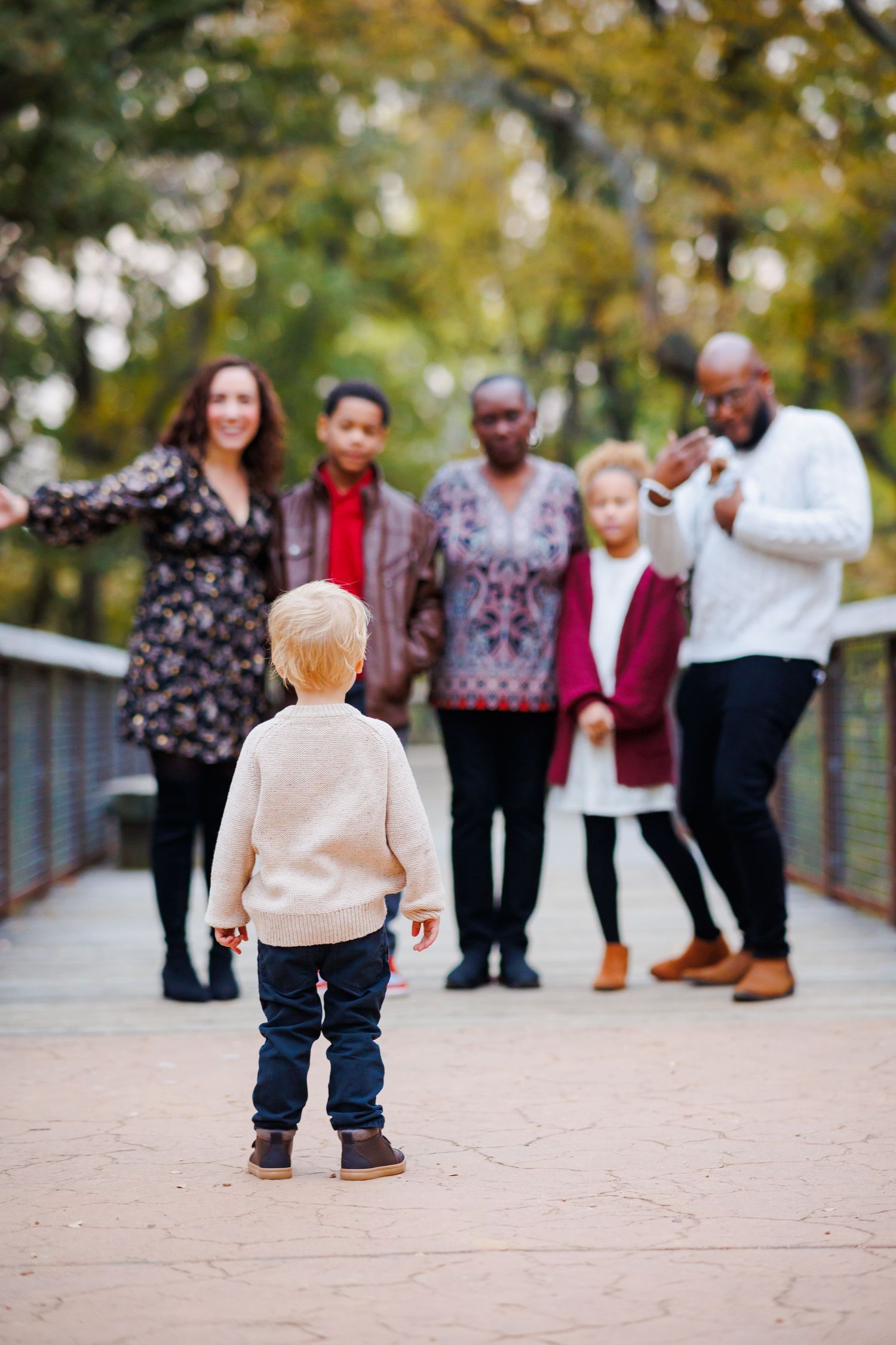 A young blonde toddler stands on a bridge facing a diverse multigenerational family in an autumn park setting.
