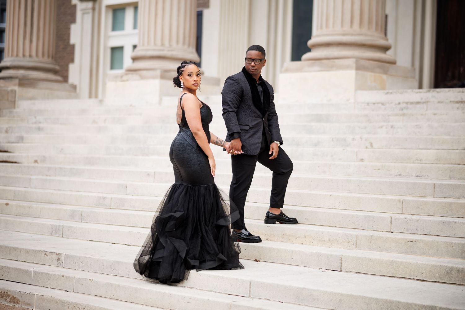 A couple in formal attire poses romantically on grand marble steps of a classic building.