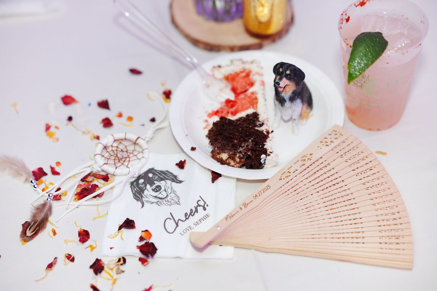 A delicate wooden fan lies beside a dessert plate with scattered rose petals and crumbs on a light surface.