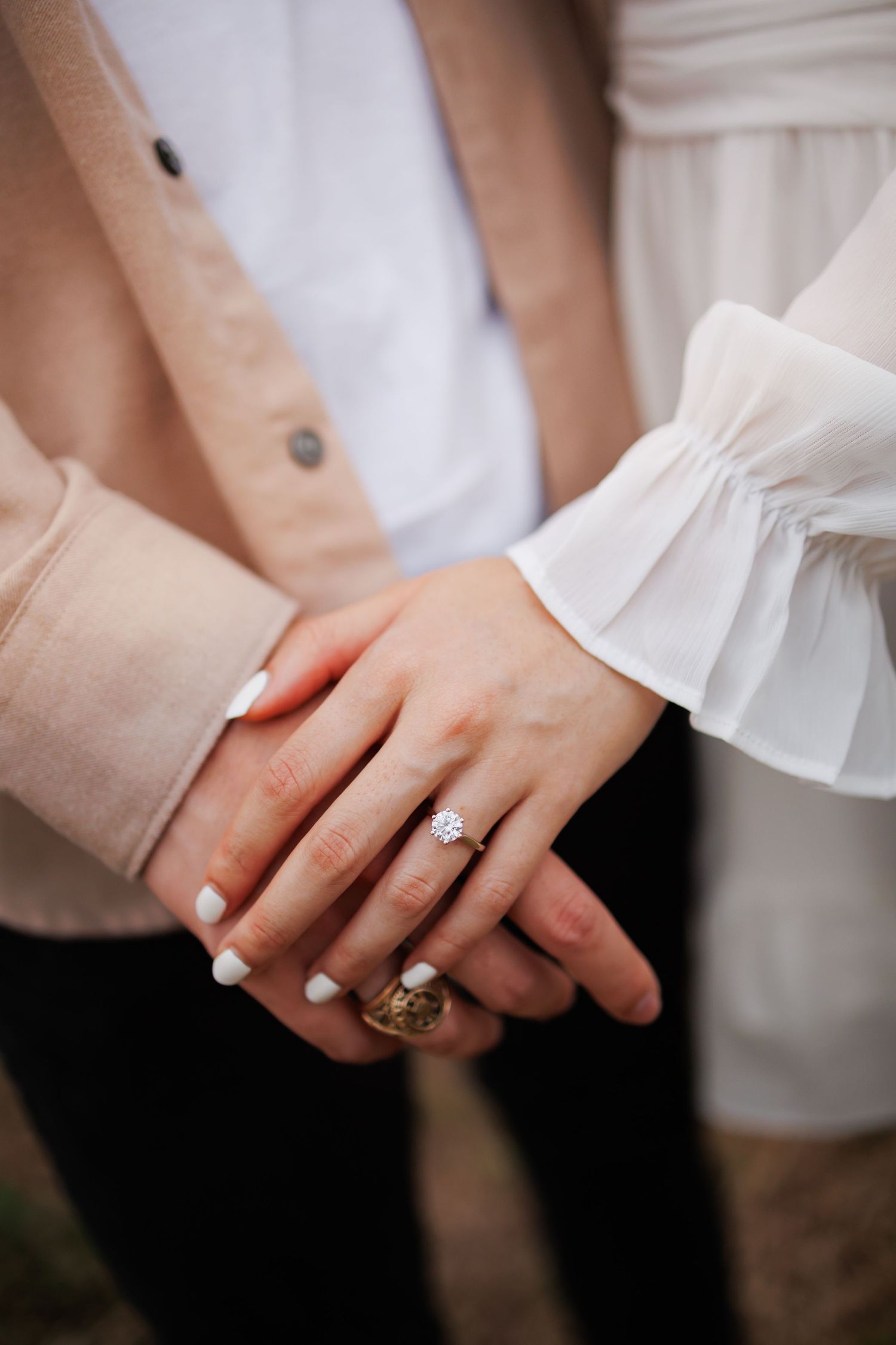 Close-up of hands wearing engagement ring and wedding band with white ruffled sleeve and beige cardigan in view.