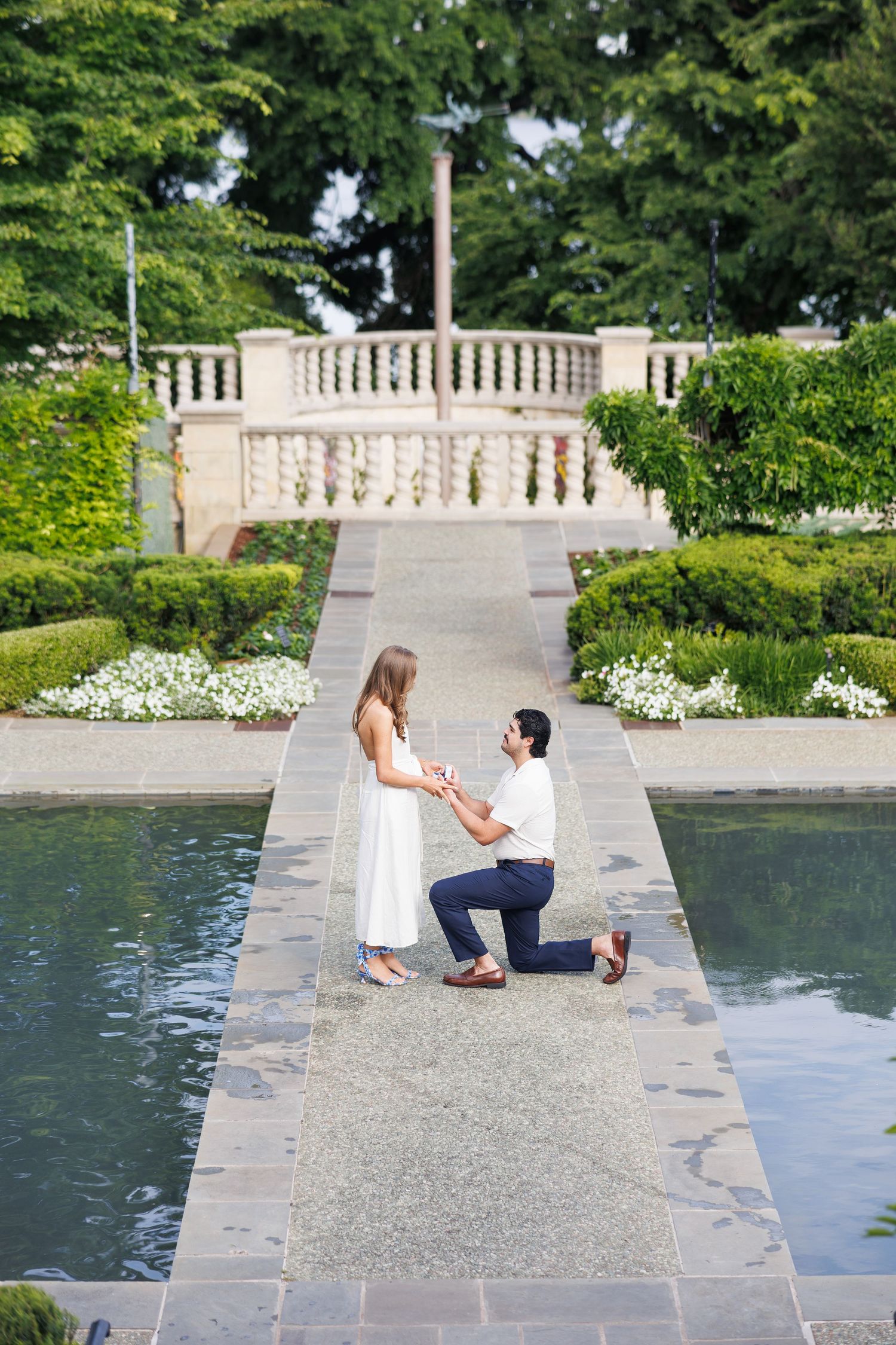 A romantic proposal sequence showing a couple on a stone terrace overlooking a reflecting pool in a garden setting.