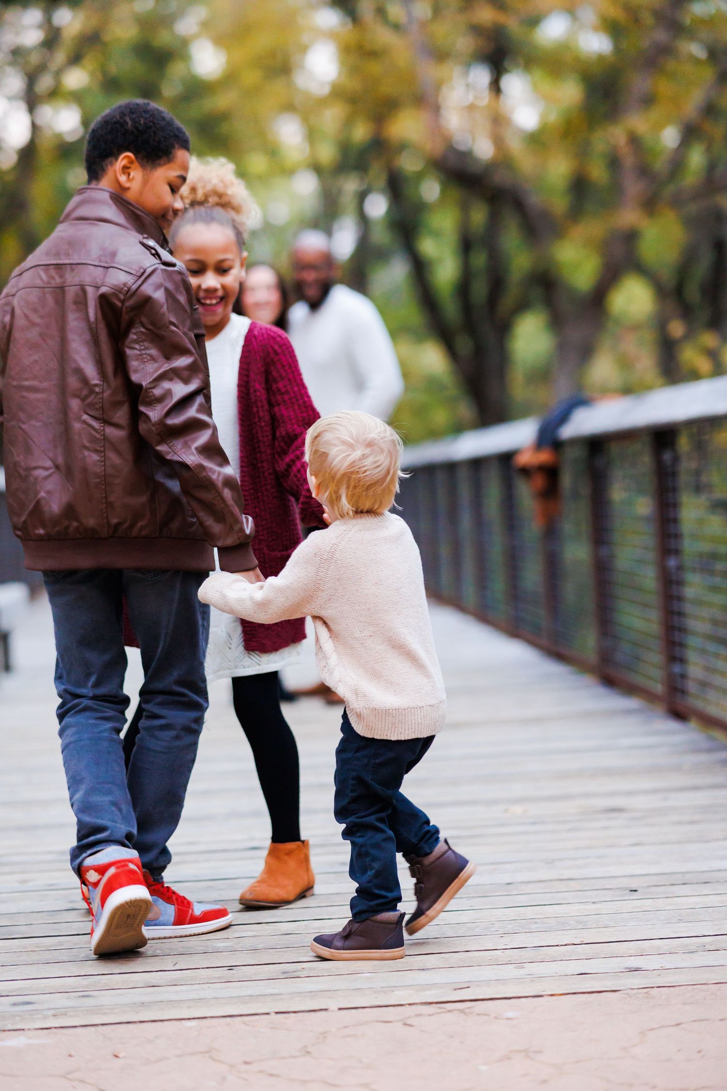 Family walking on a wooden bridge in an autumn park, with children holding hands with parents outdoors.