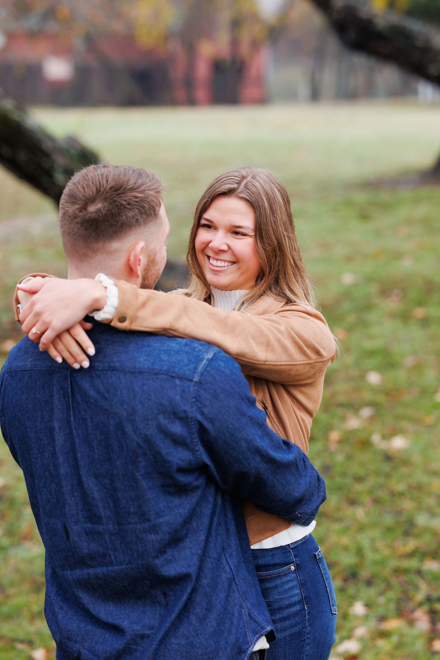 Smiling woman in tan jacket embraces man in denim shirt outdoors in a park during autumn.