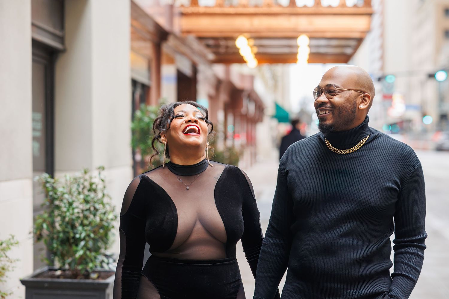 A joyful couple wearing black sweaters share a candid laughing moment together on a city street.
