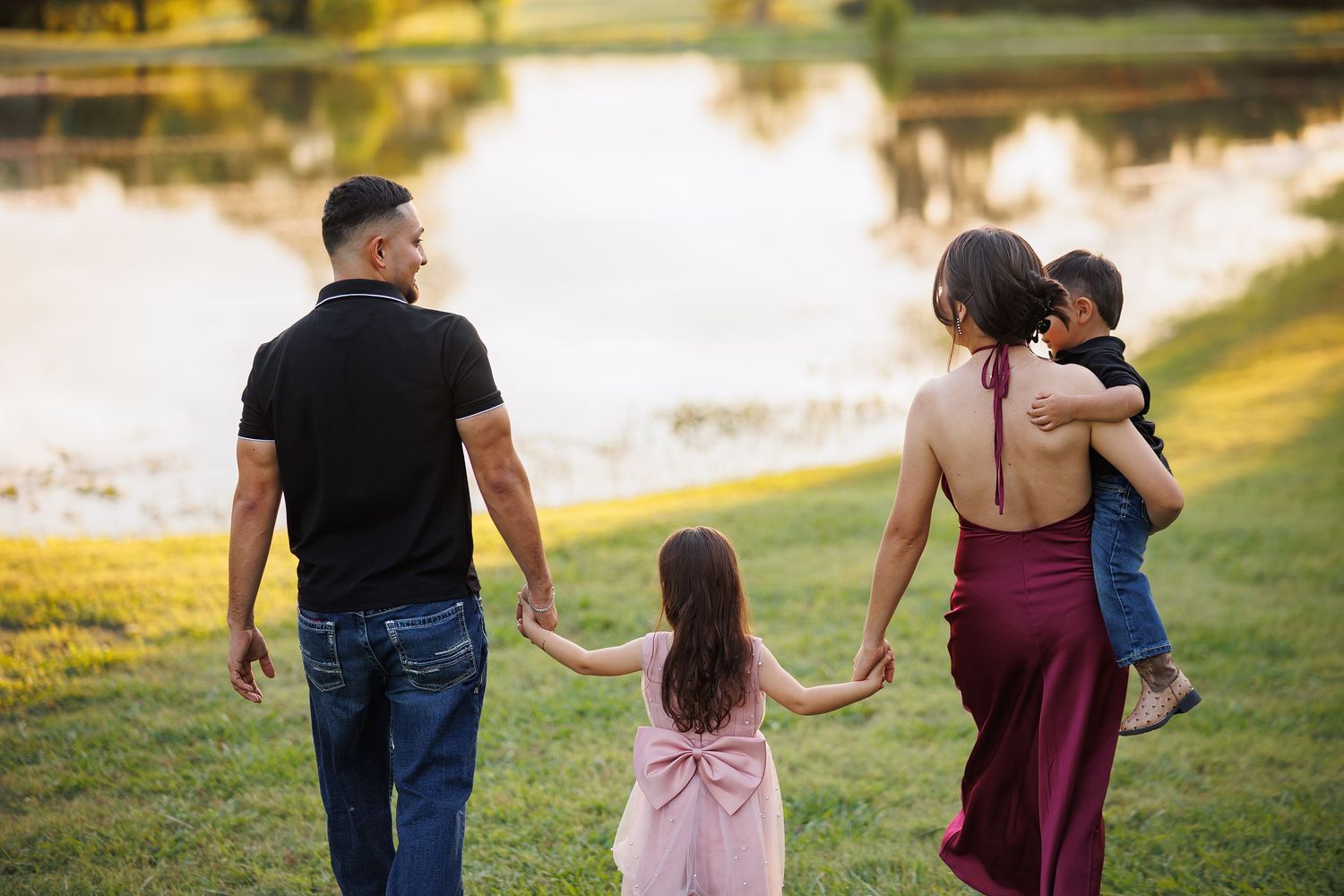 Family of four walking hand-in-hand toward a serene pond in a lush green park at golden hour.
