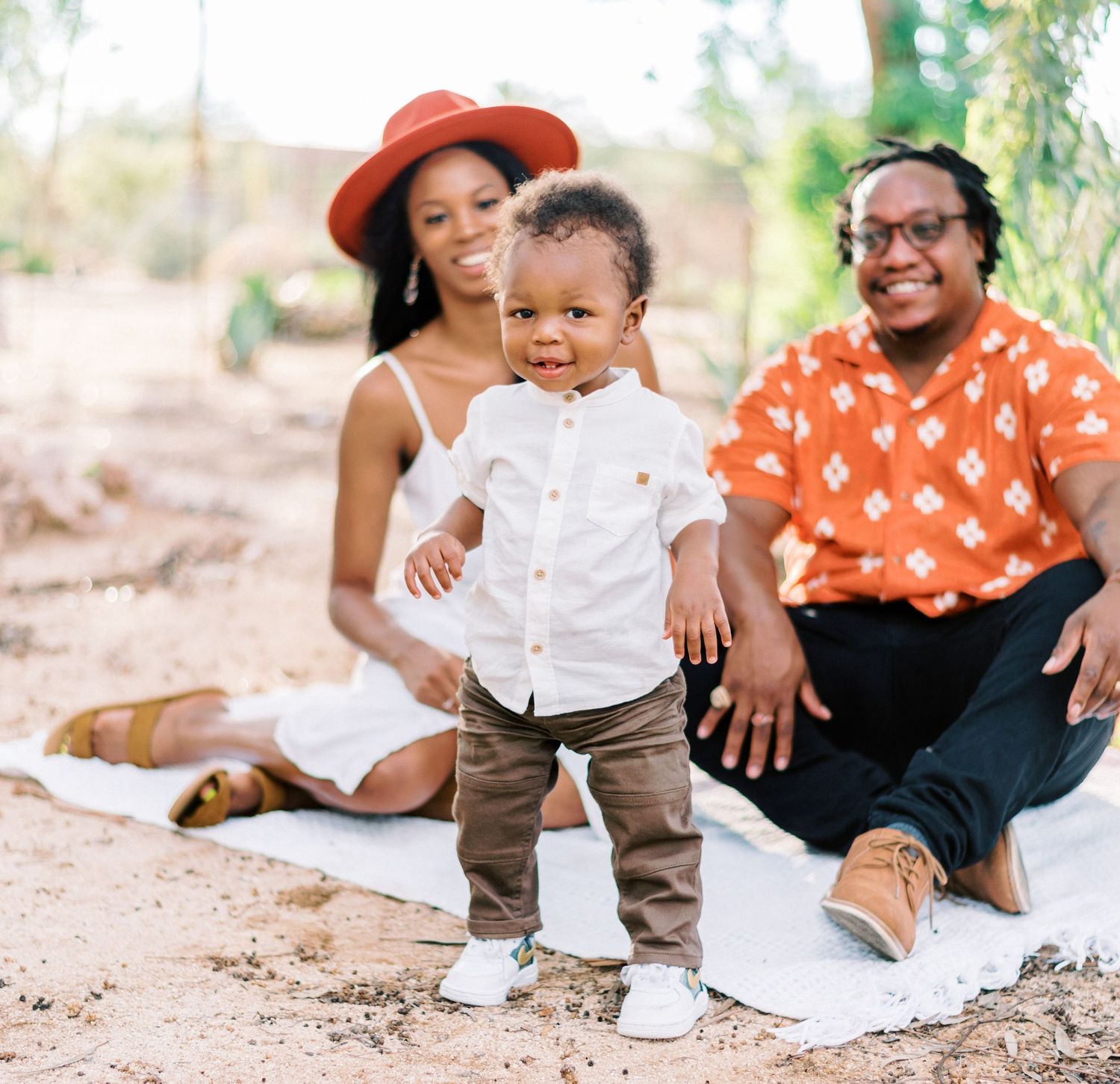 A family sits together on a white blanket outdoors during a casual photo session in natural lighting.