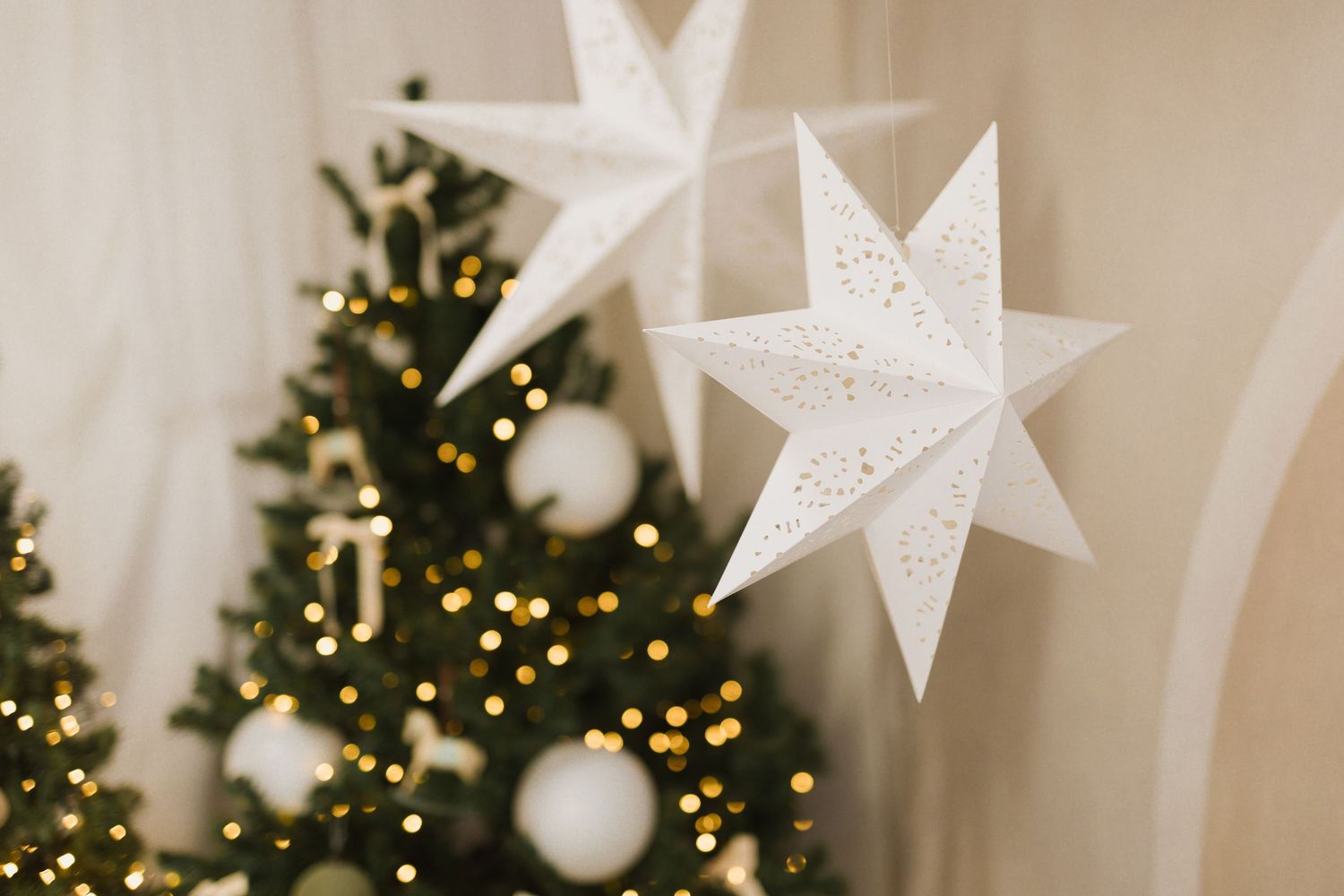 White paper star ornaments hanging near a decorated Christmas tree with golden lights and white ball ornaments.