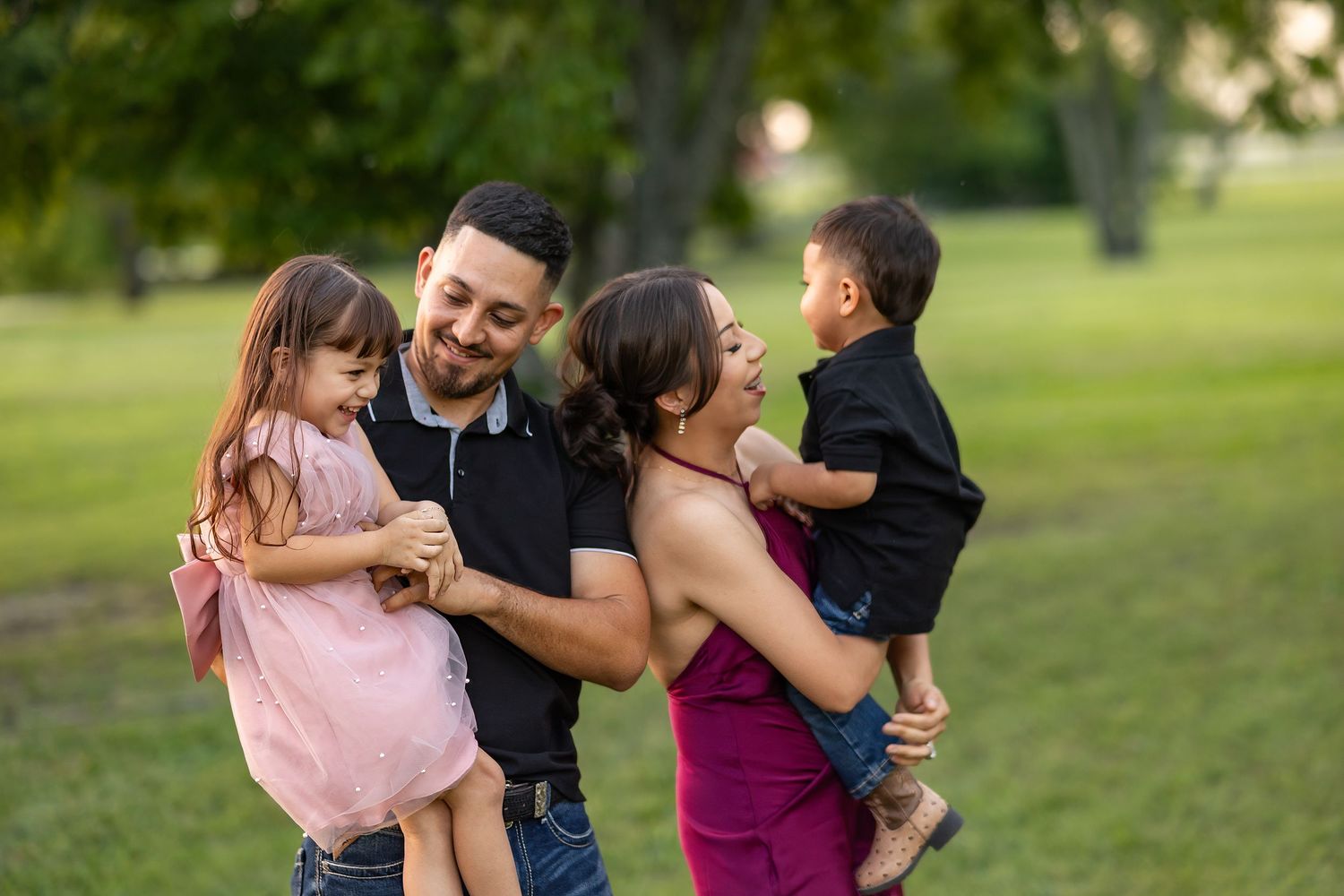Happy family of four laughing together outdoors in a green park, parents holding young children.