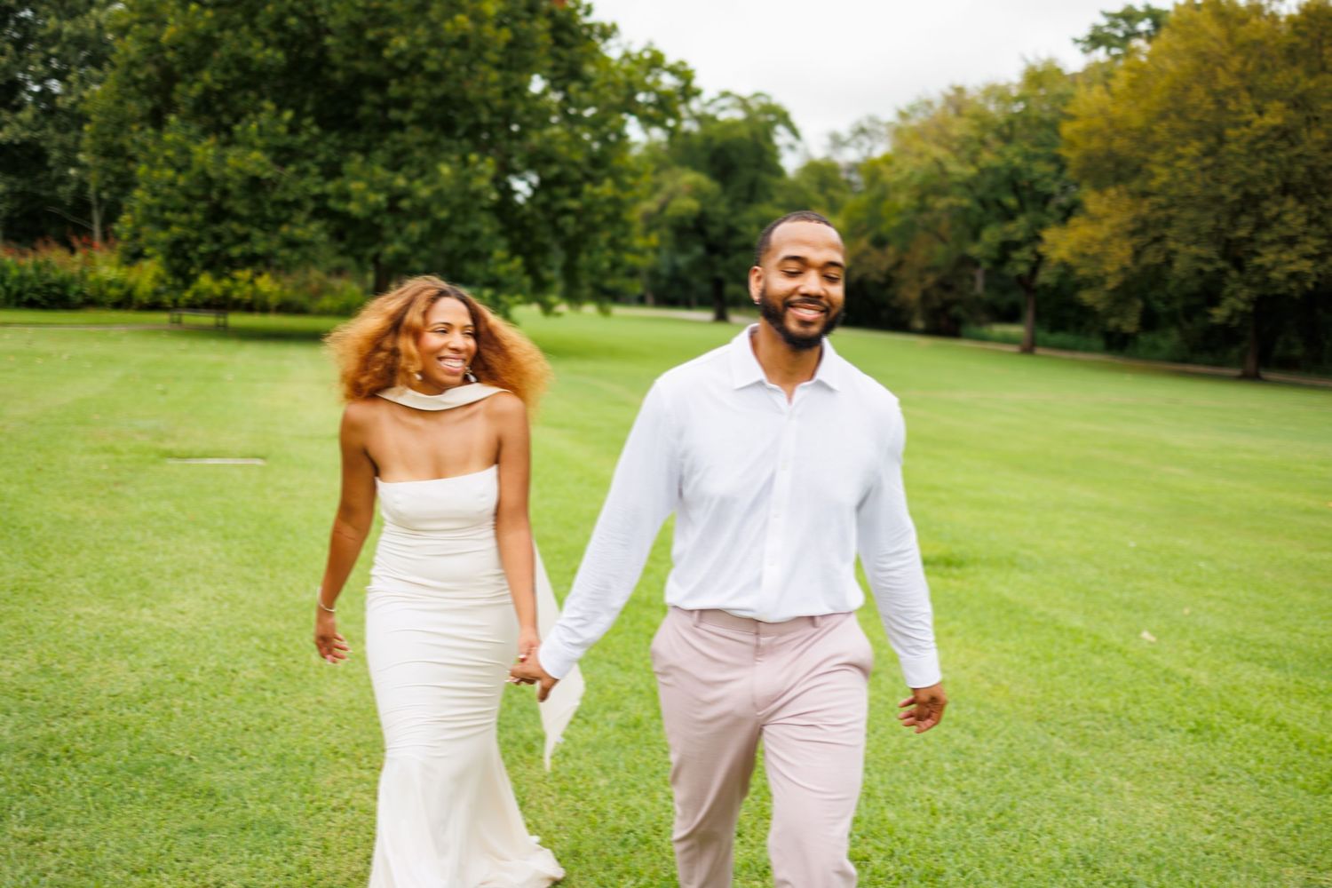 A couple in wedding attire walks hand in hand through a lush green park on a sunny autumn day.