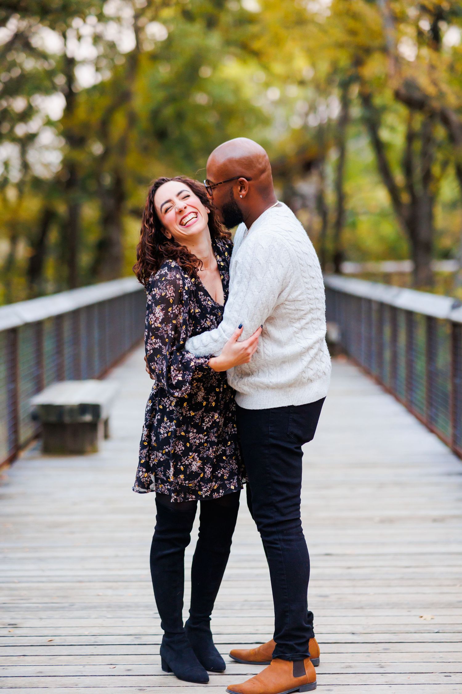 A joyful interracial couple embraces and laughs together on a wooden bridge surrounded by autumn foliage.