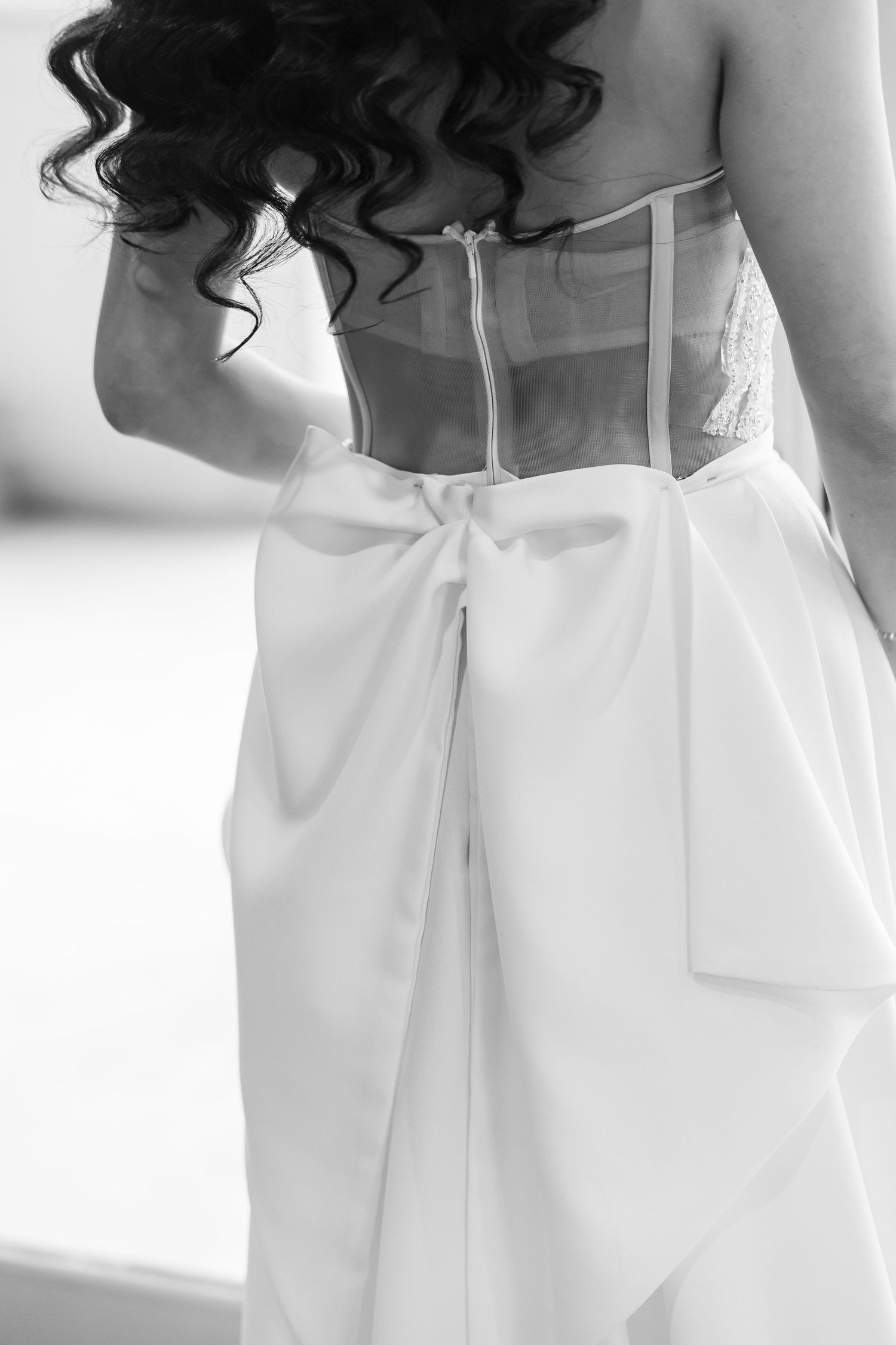 Black and white photo showing the back detail of a wedding dress with corset bodice and flowing white fabric bow.