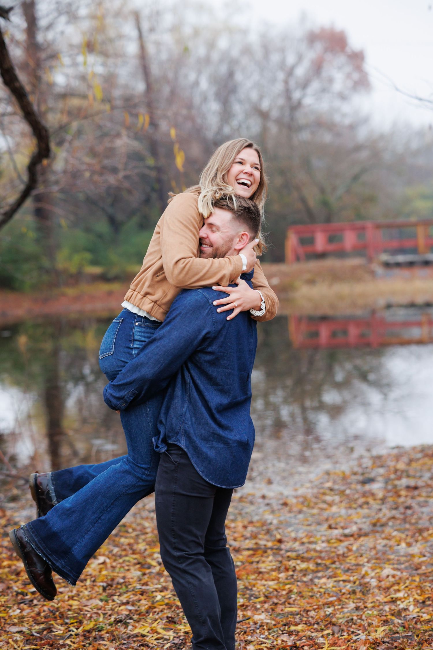 A couple shares a joyful embrace in a park during autumn with fallen leaves and a red bridge in the background.