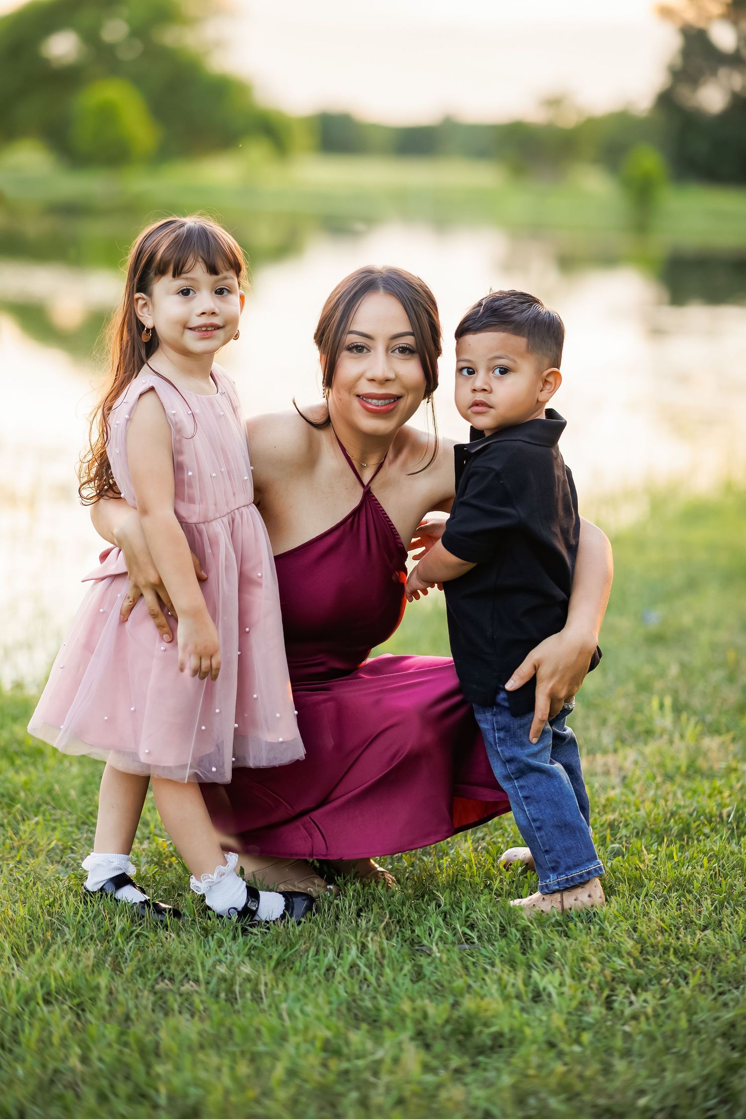 A mother in a burgundy dress crouches with her young daughter in pink and toddler son in black shirt outdoors.