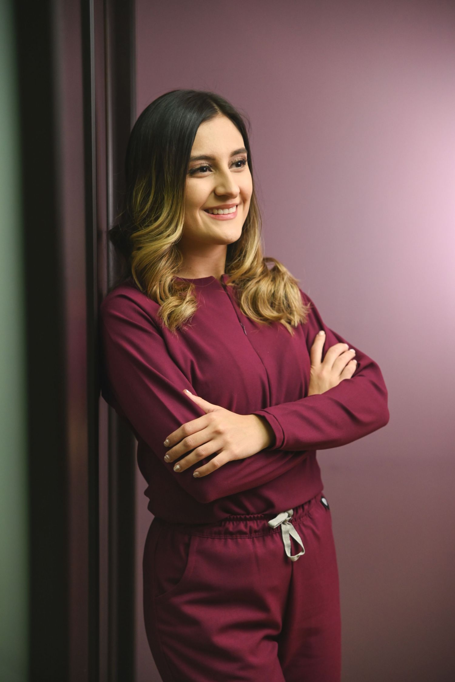 Professional portrait of someone in a burgundy sweatsuit posing with arms crossed and smiling against a pink background.