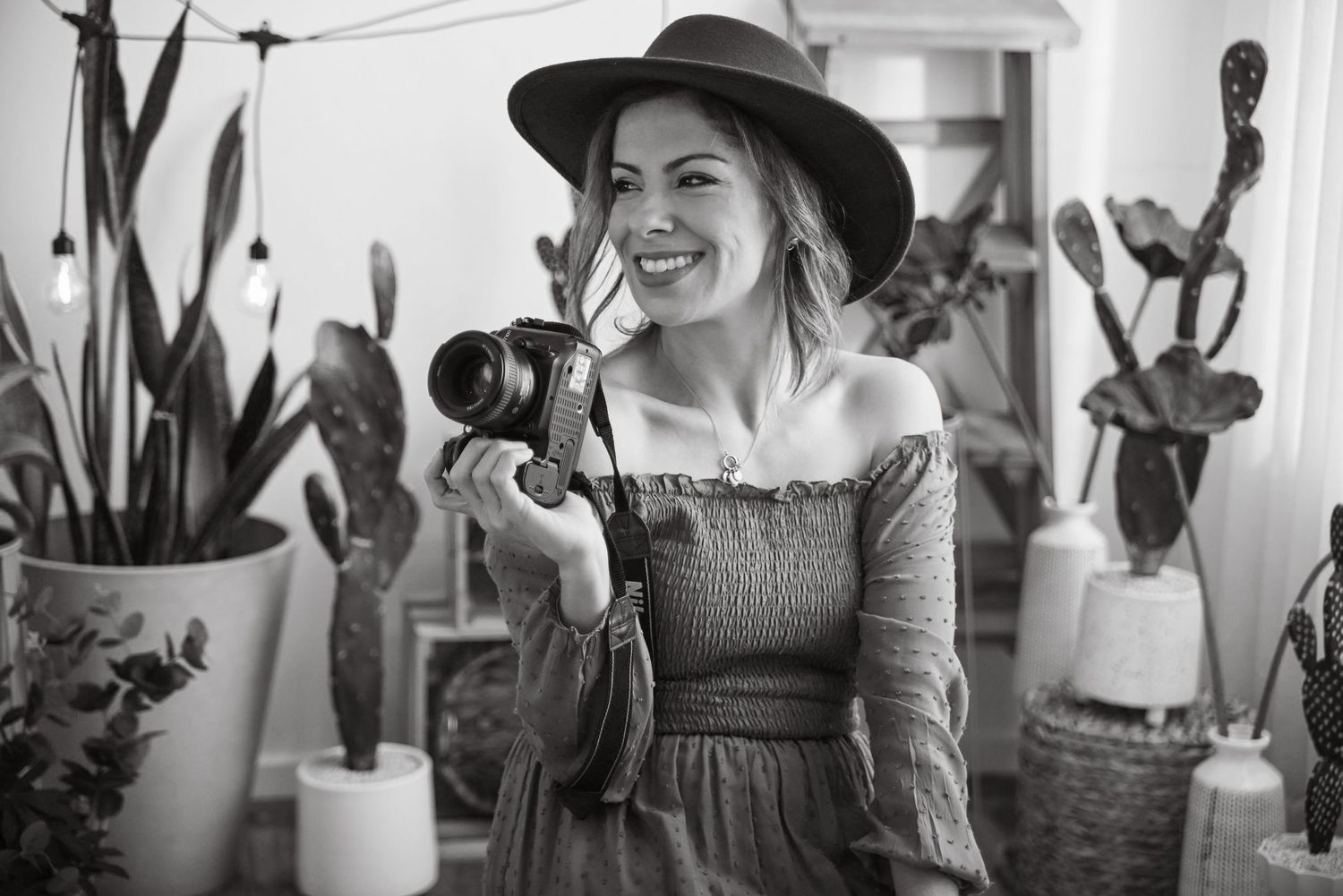 A photographer in a wide-brimmed hat and dress smiles while holding a camera in a room with potted plants.