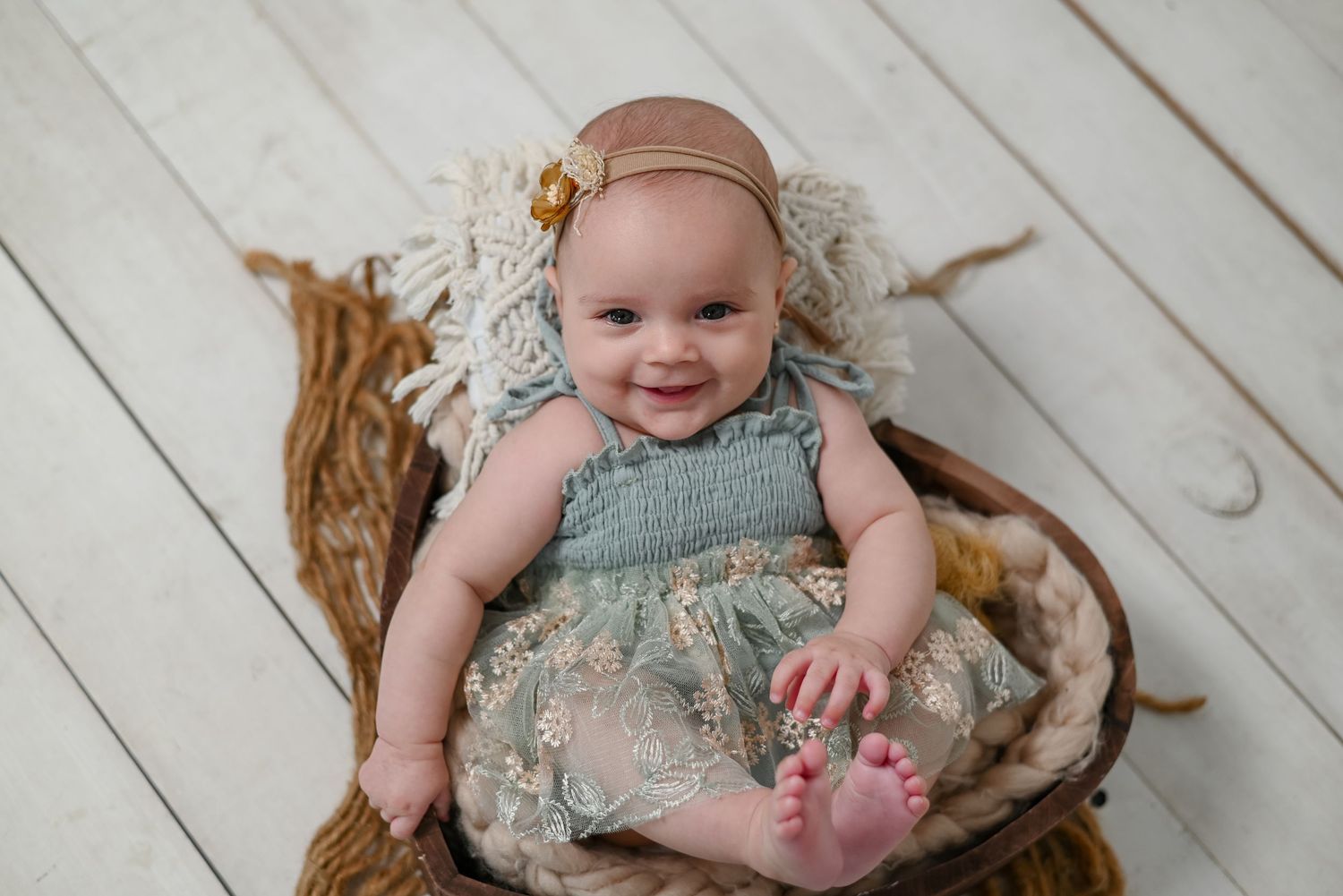 Baby wearing blue floral dress smiles while sitting on textured brown blanket against white wooden floor.