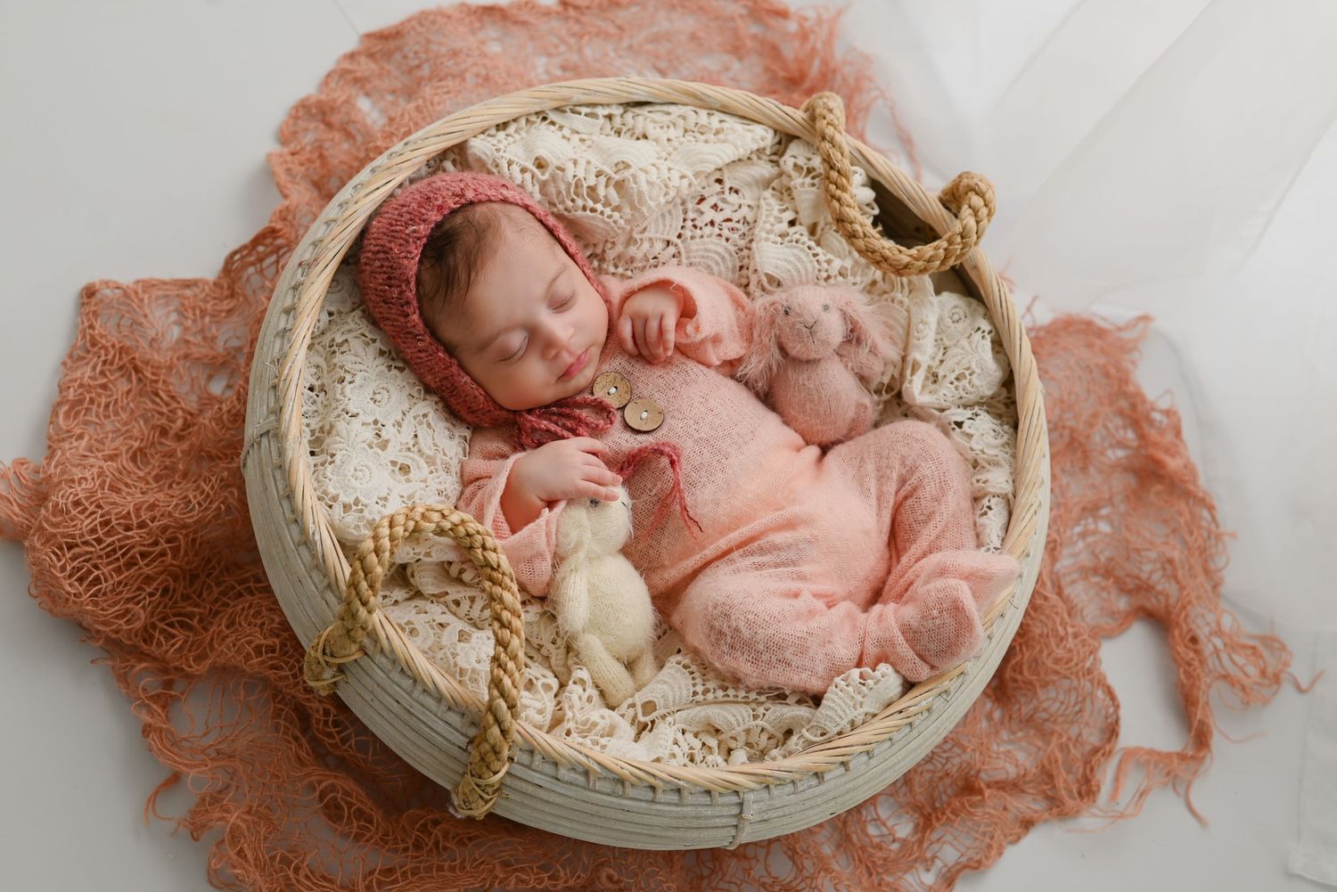 Sleeping baby in pink outfit lies peacefully in round white basket with peach-colored fabric backdrop.