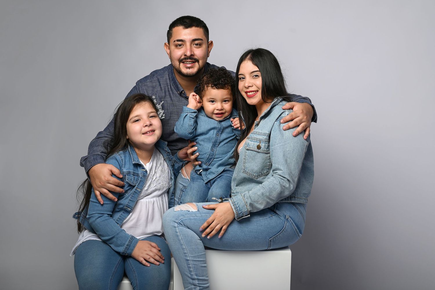 Family in matching denim outfits smiling together against grey background during studio photoshoot.
