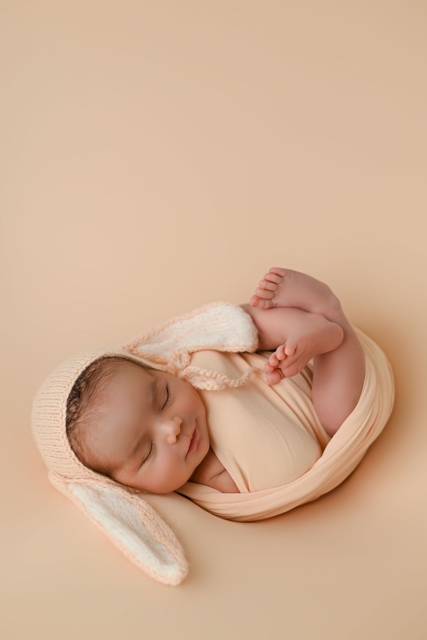 Sleeping newborn wrapped in cream fabric with knitted bunny ears.