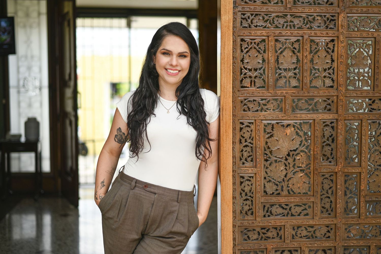 Beautiful woman in white top and brown pants stands near ornate wooden carved door in elegant interior setting.
