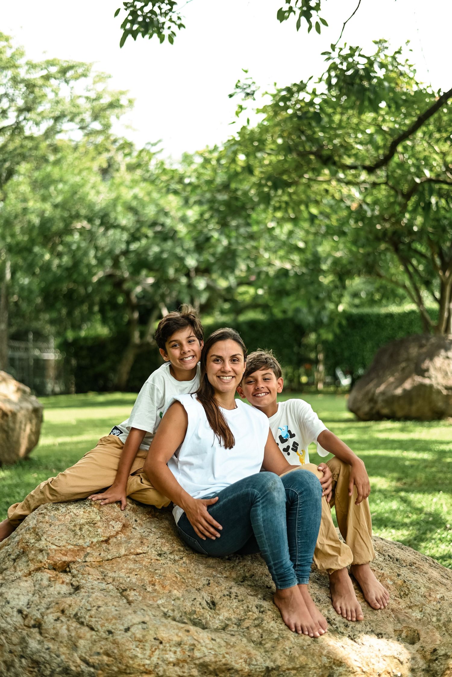 A family sits together on a large boulder in a sunlit park surrounded by lush green trees.