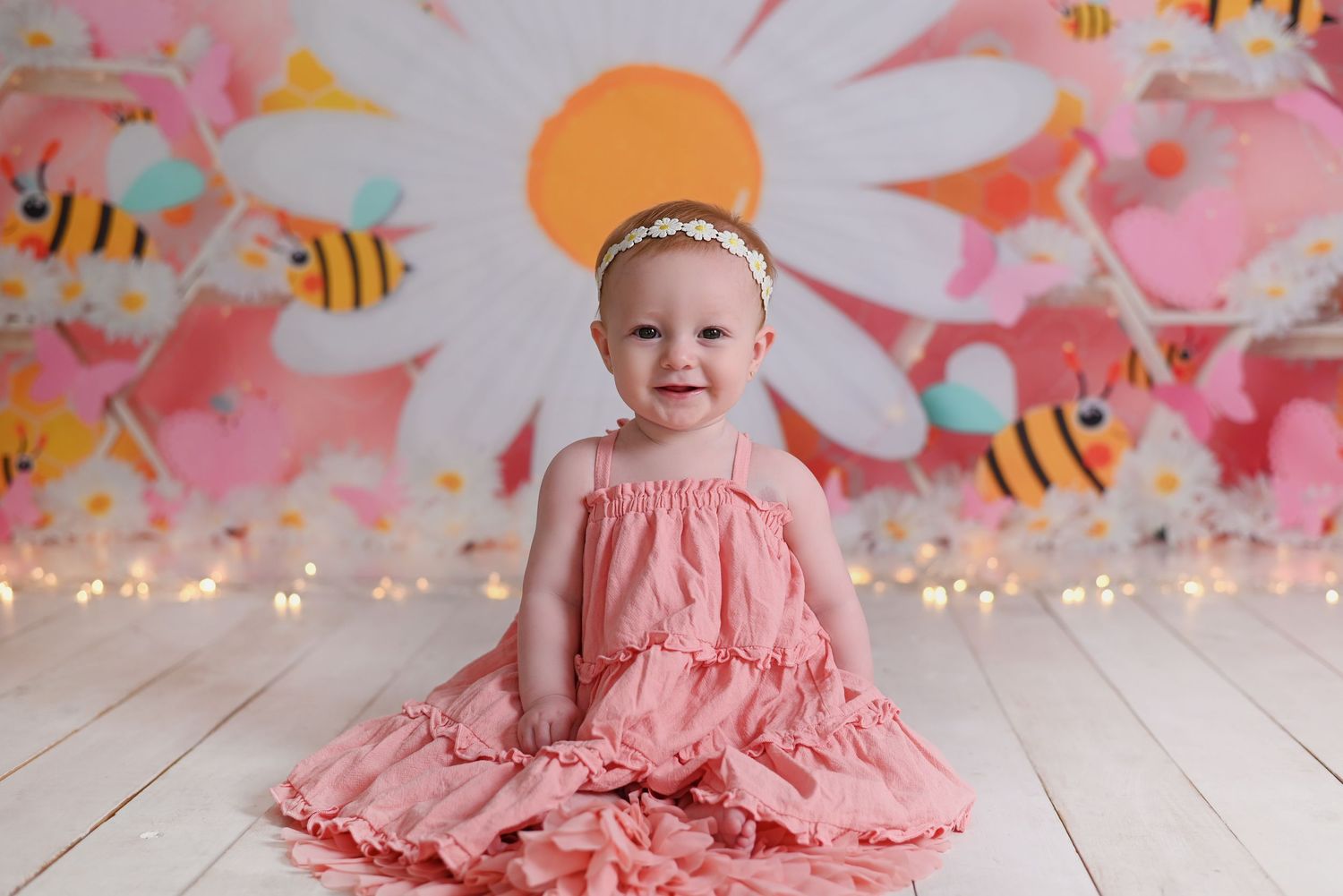 Young child in pink ruffled dress sits on white wooden floor against pastel backdrop with cartoon bee decorations.