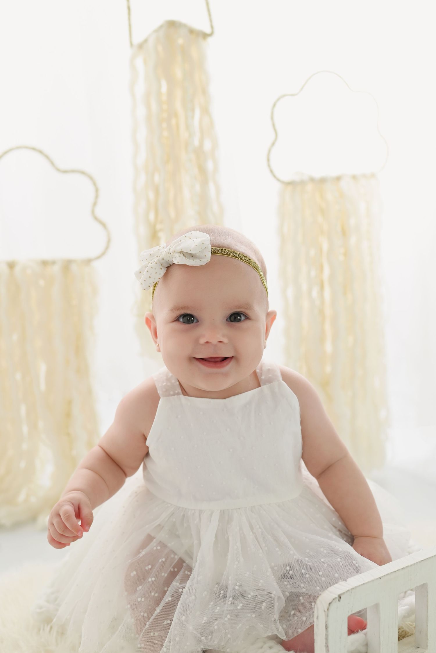 Baby wearing white tulle dress and headband sits smiling in front of light curtains in bright natural setting.
