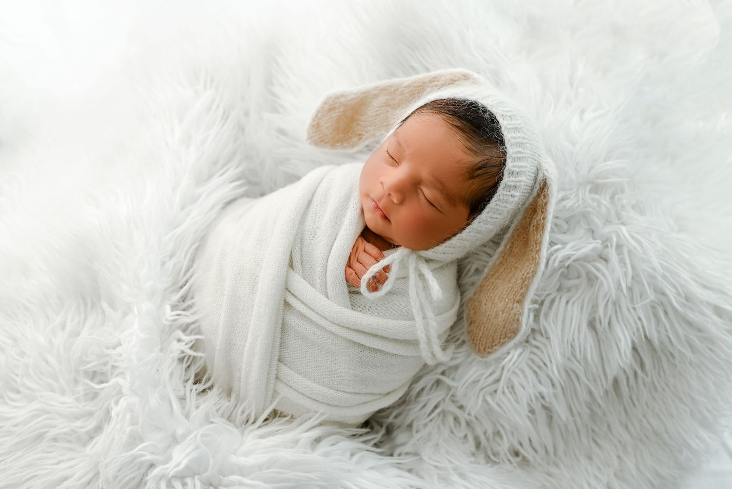 Newborn baby yawning while wrapped in white blanket on fur backdrop.