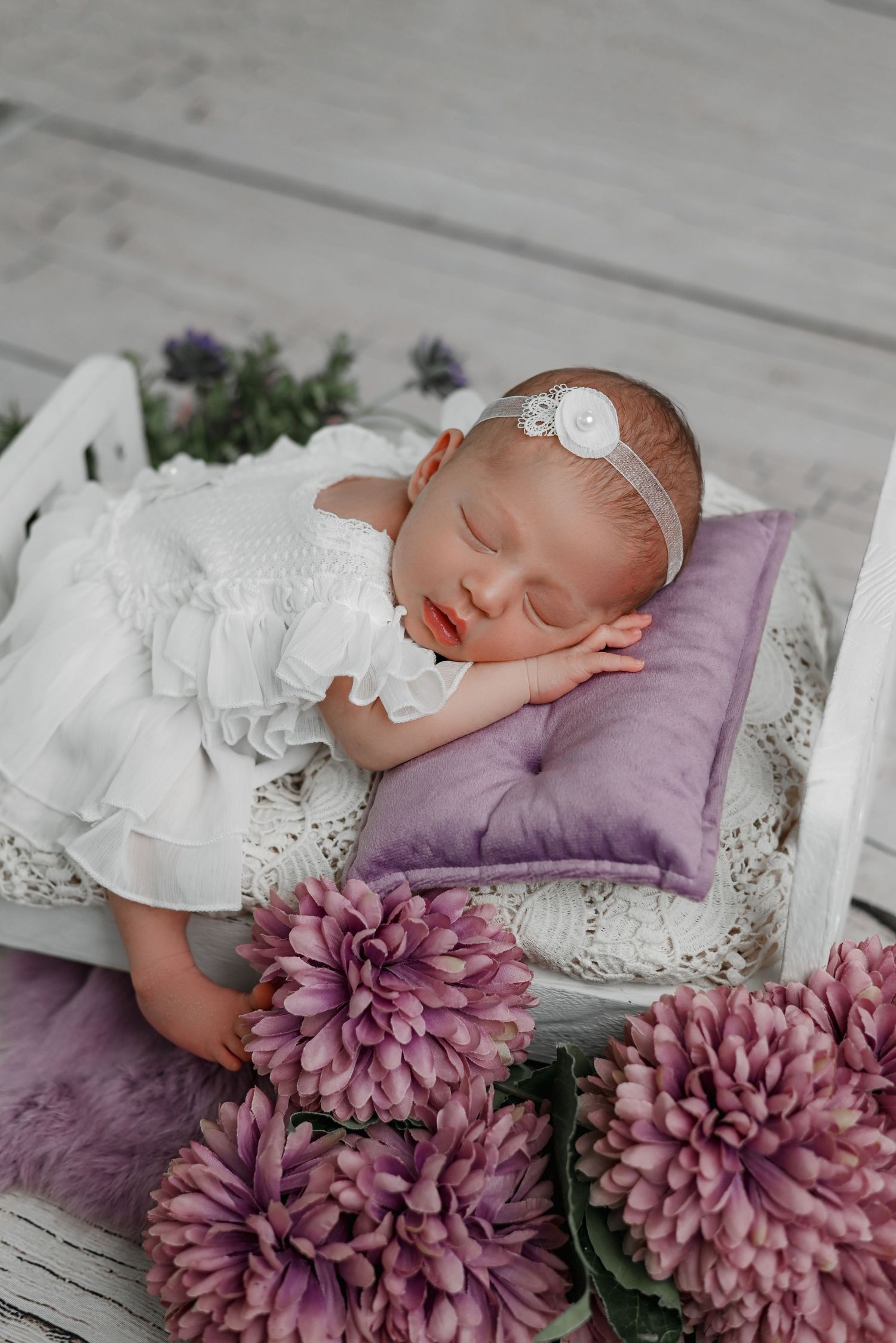 Newborn on purple pillow with white dress surrounded by mauve flowers.