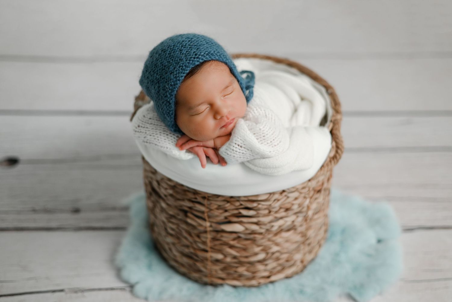 Sleeping newborn in teal knit hat nestled in woven basket.