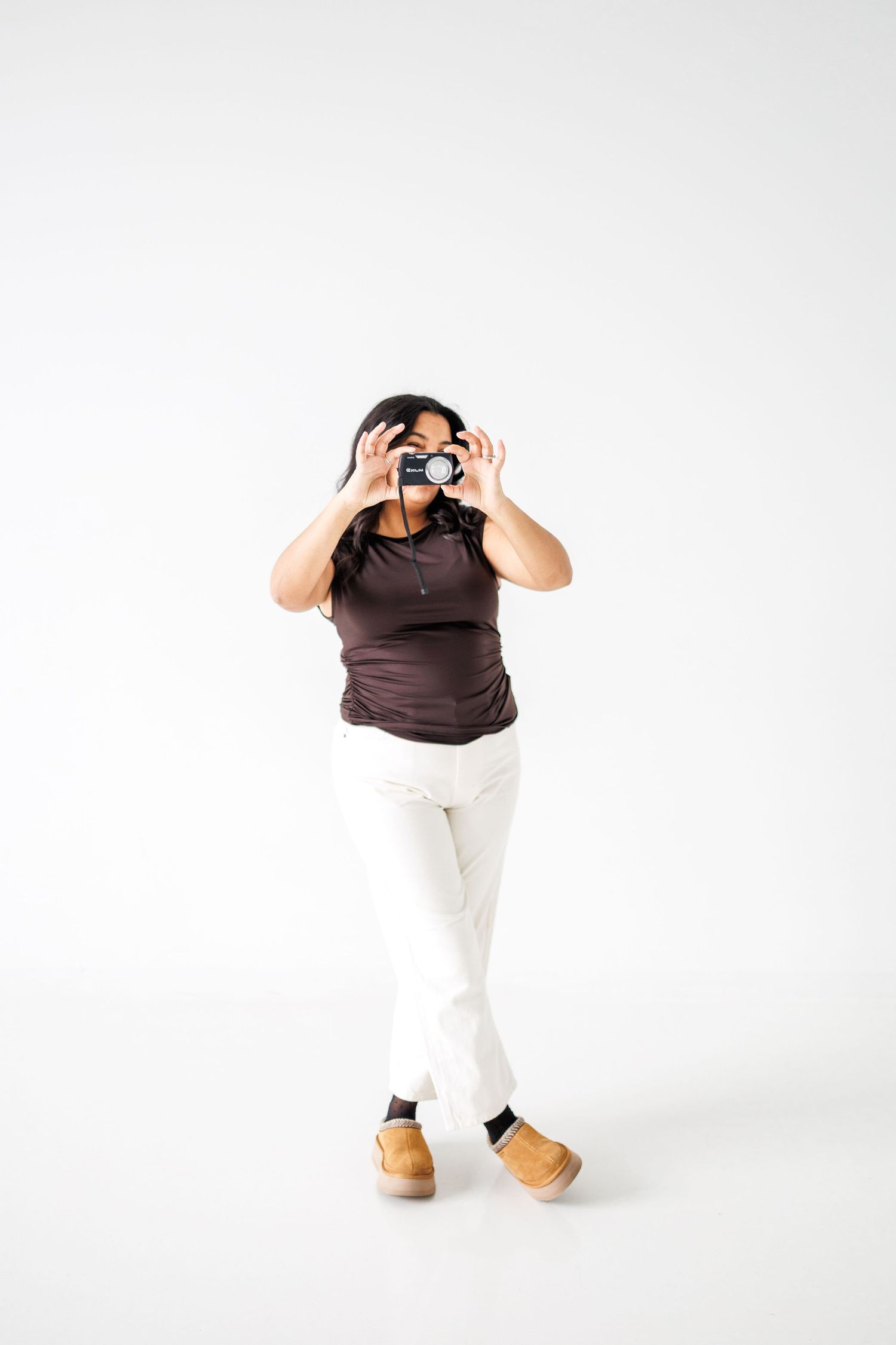 Person in brown shirt and white pants stands against white background with hands raised to head in expressive pose.
