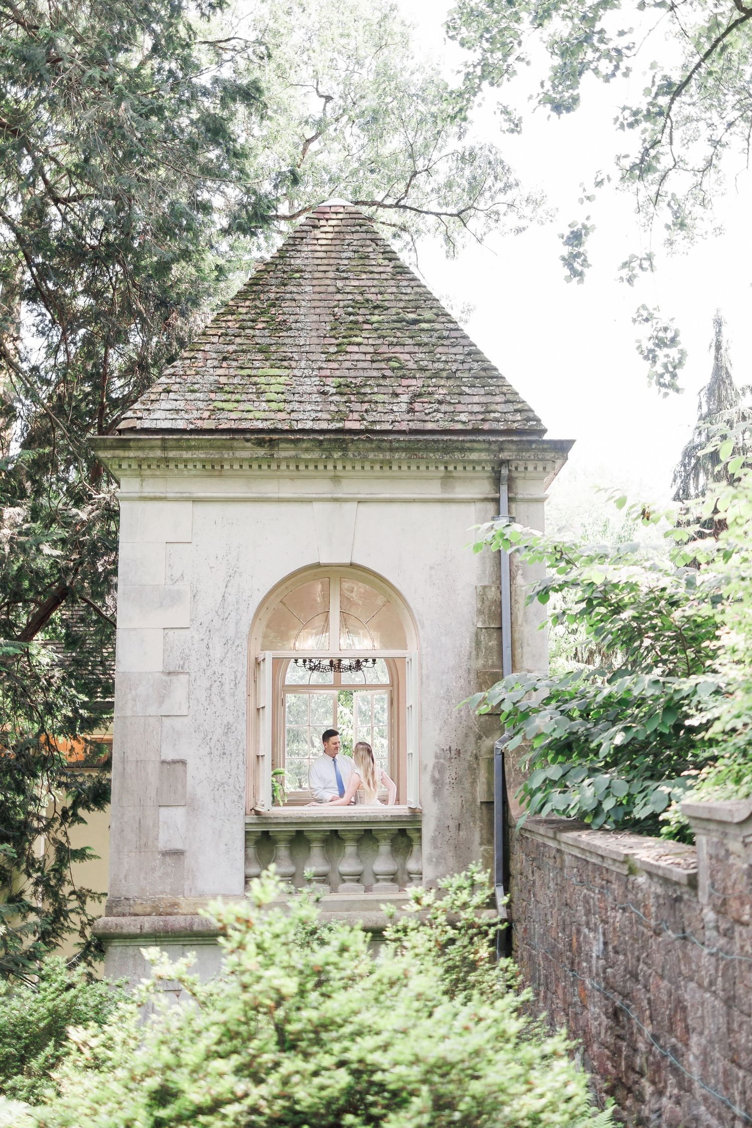 A series of stone garden pavilions with arched openings connected by a rustic brick wall in a lush garden setting.