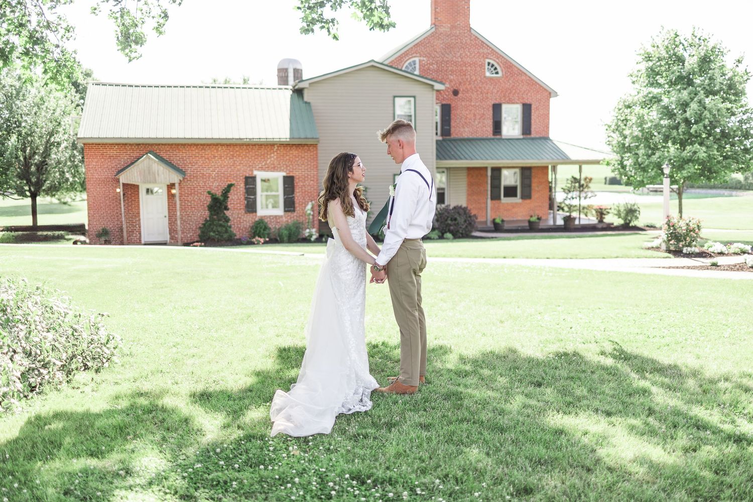 A person in a white dress walks through a sunny garden setting near brick buildings.